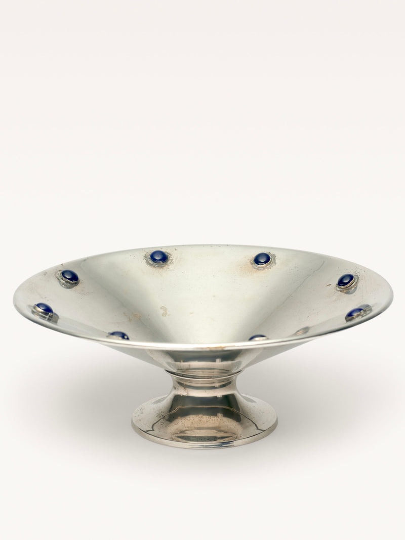 The "Silver Plated Bowl with Blue Stones" by Galerie MLS is a wide, shallow pedestal bowl featuring blue stones around the rim. It has a reflective surface, stands on a rounded base, and shows slight signs of use that enhance its elegance.