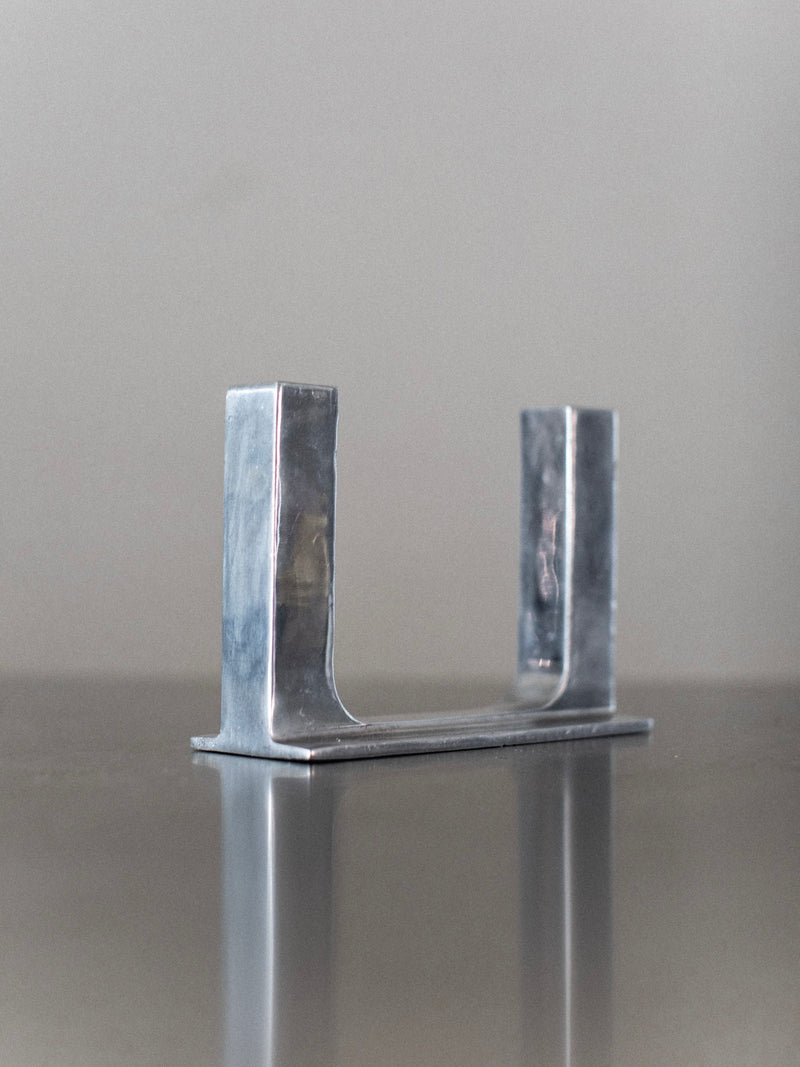 The Chrome Candleholder by Out For Lunch, a minimalist U-shaped steel piece with a subtle polished silver finish, is placed on a reflective surface. A plain, muted background allows this elegant object to take center stage, its metal catching the light with a gentle shine.
