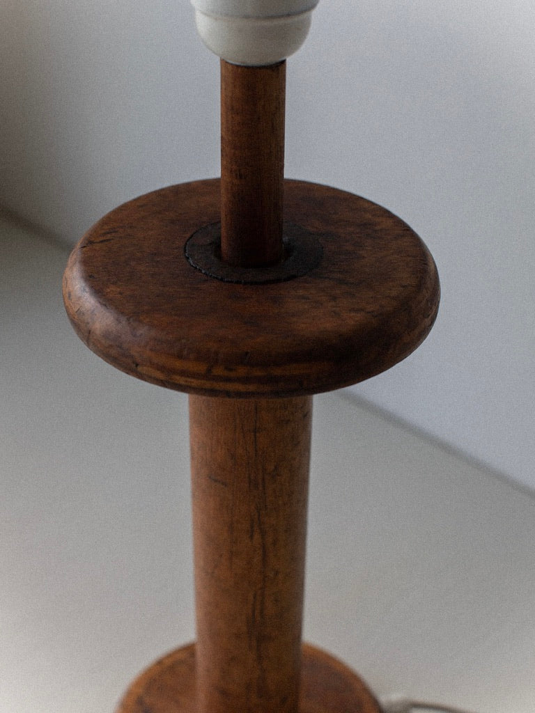 A close-up of the Midcentury Table Lamp by Out For Lunch. The cylindrical wooden handle base showcases a round, flat platform near the top and bottom. The wood offers a natural finish with a slightly worn appearance, and the light socket is partially visible at the top of the image.