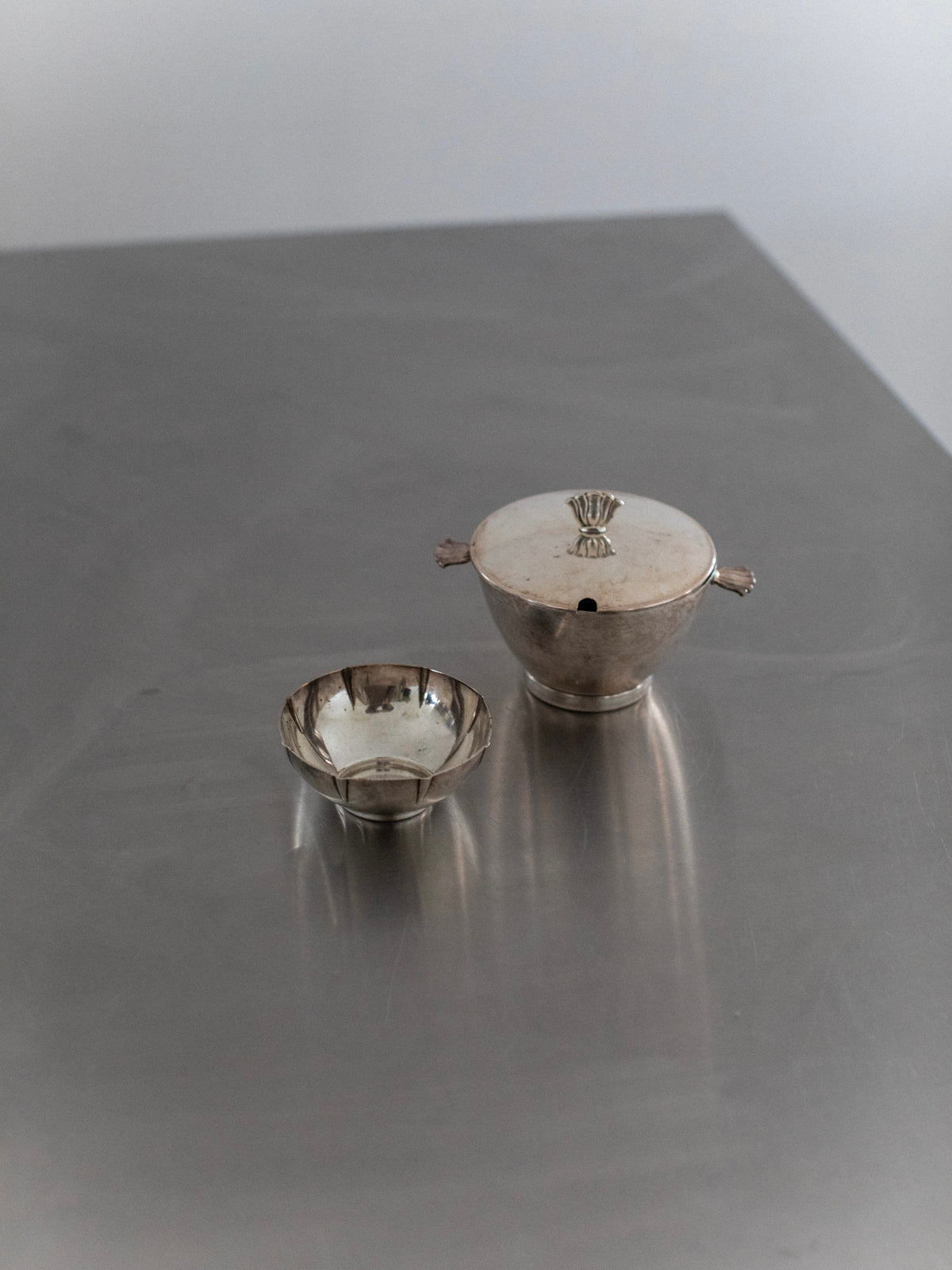 A touch of vintage glamour is added to the plain, softly lit background with an Out For Lunch Art Deco lidded bowl in a small silver design, featuring handles and a decorative finial, placed on a reflective steel surface next to its matching counterpart.