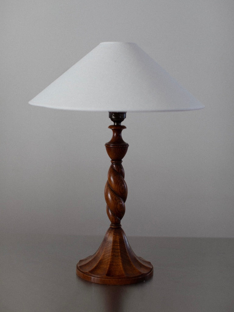 The Early 1900's Table Lamp by Out For Lunch features a wooden base with intricate carvings in a rich brown finish and a cone-shaped white lampshade. It sits on a flat surface against a plain, light background, adding a touch of vintage decor to any room.