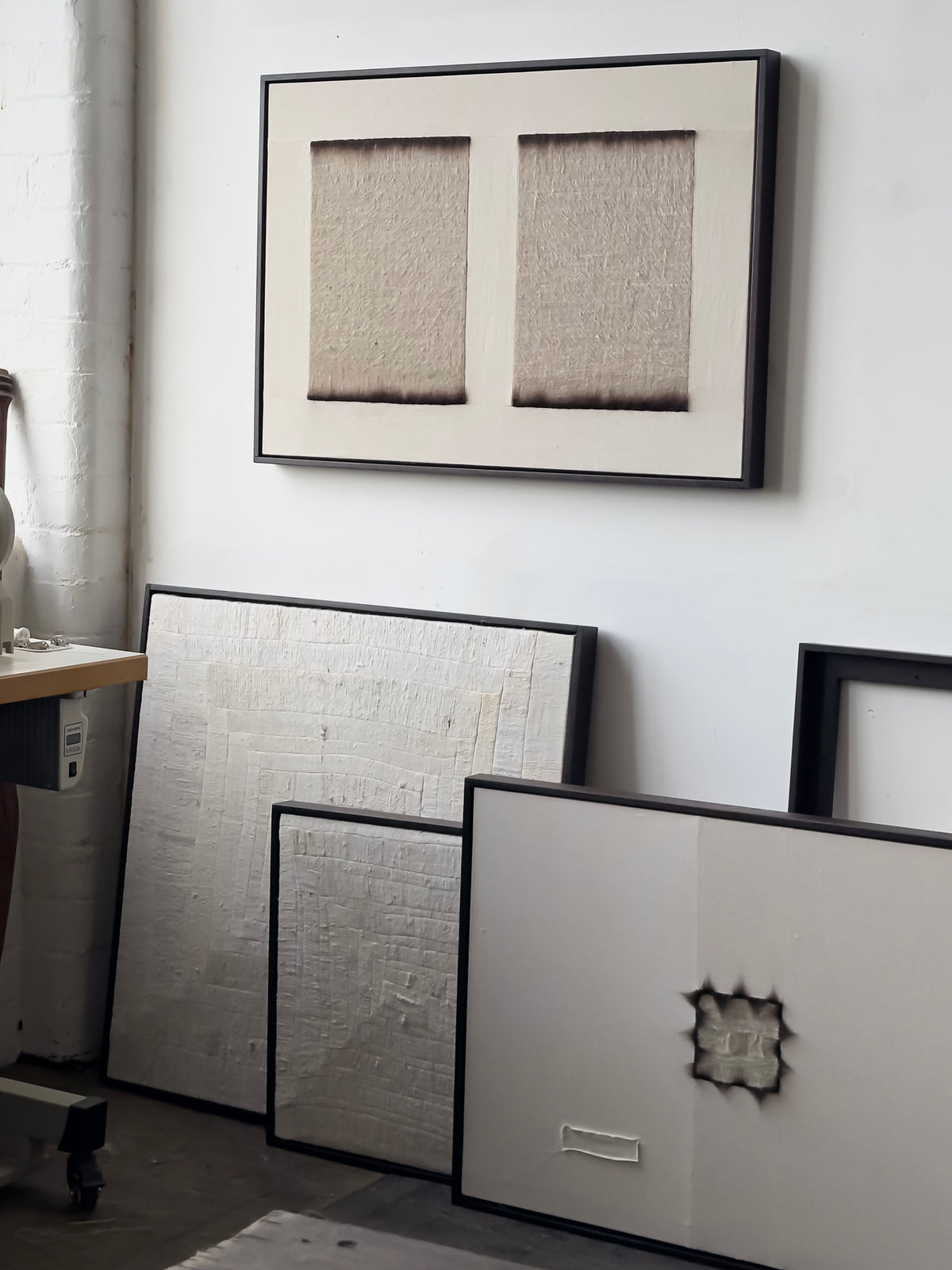 Bec Kirby's 'To Hold', 2025 framed abstract artworks adorn a white wall. One piece depicting two textured rectangles is hung, while others, including one with a starburst indentation, lean on the floor. To the left, a sewing machine on solid oak whispers tales of creation and craft.