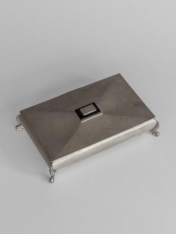 Introducing the Art Deco Metal Box by Spigel, a rectangular piece of Scandinavian craftsmanship. This metallic box is adorned with a small square handle on top and supported by four ornate feet. Its surface features a subtly domed design, capturing the essence of the Art Deco style, set against a plain and neutral background.