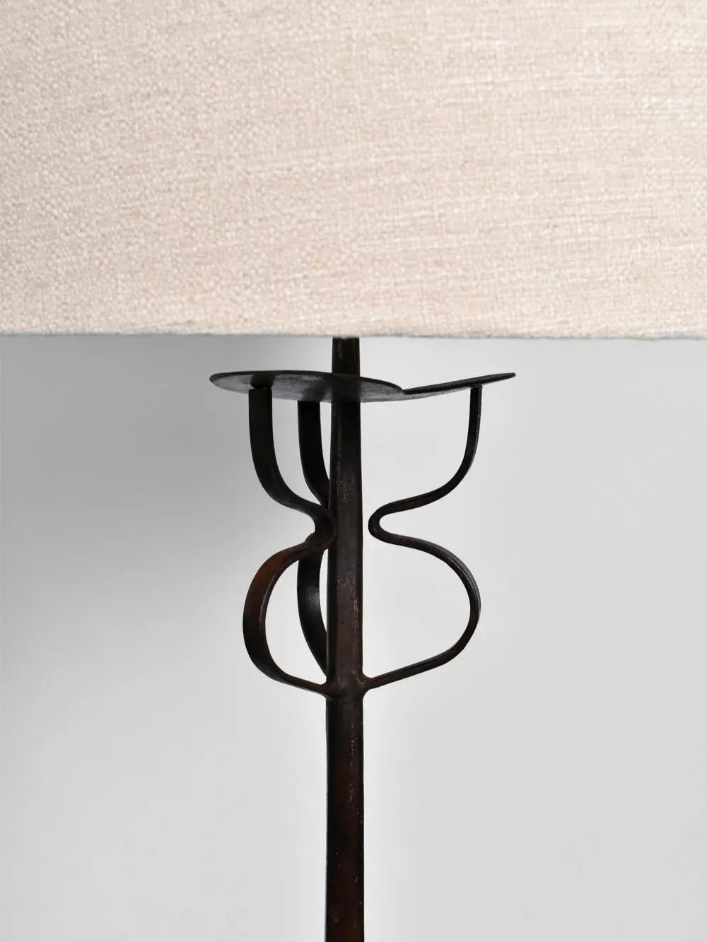 A close-up of Barracuda Interiors' Tripod Iron Floor Lamp, 1960s, showcases its sculptural design with a beige lampshade. The lamp features a wrought iron base with abstract, curved shapes inspired by 1960s France, offering a modern artistic touch. The background is plain and light-colored.