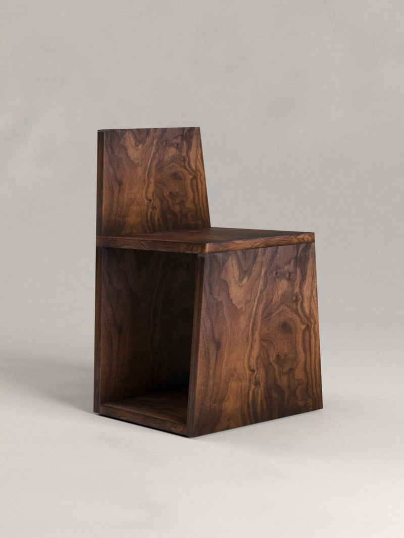 Introducing Siete Studio's 'Totem' Chair: a modern, minimalist piece crafted from solid walnut. Its geometric design with a backrest and flat seat sits on an angled, hollow base. The rich dark finish showcases grain patterns, creating a totemic effect against its neutral gray backdrop.