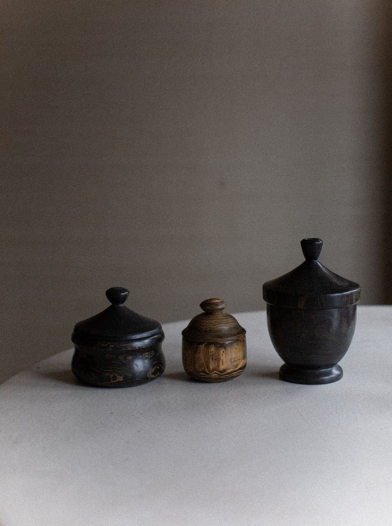 Three small wooden containers with lids are displayed on a white surface against a plain background. The container on the left and right, showcasing rustic elegance, are dark wood with a weathered texture, while the middle container is a lighter brown with intricate carvings that highlight natural wood grain. This is the Set of Three Boxes by Out For Lunch.