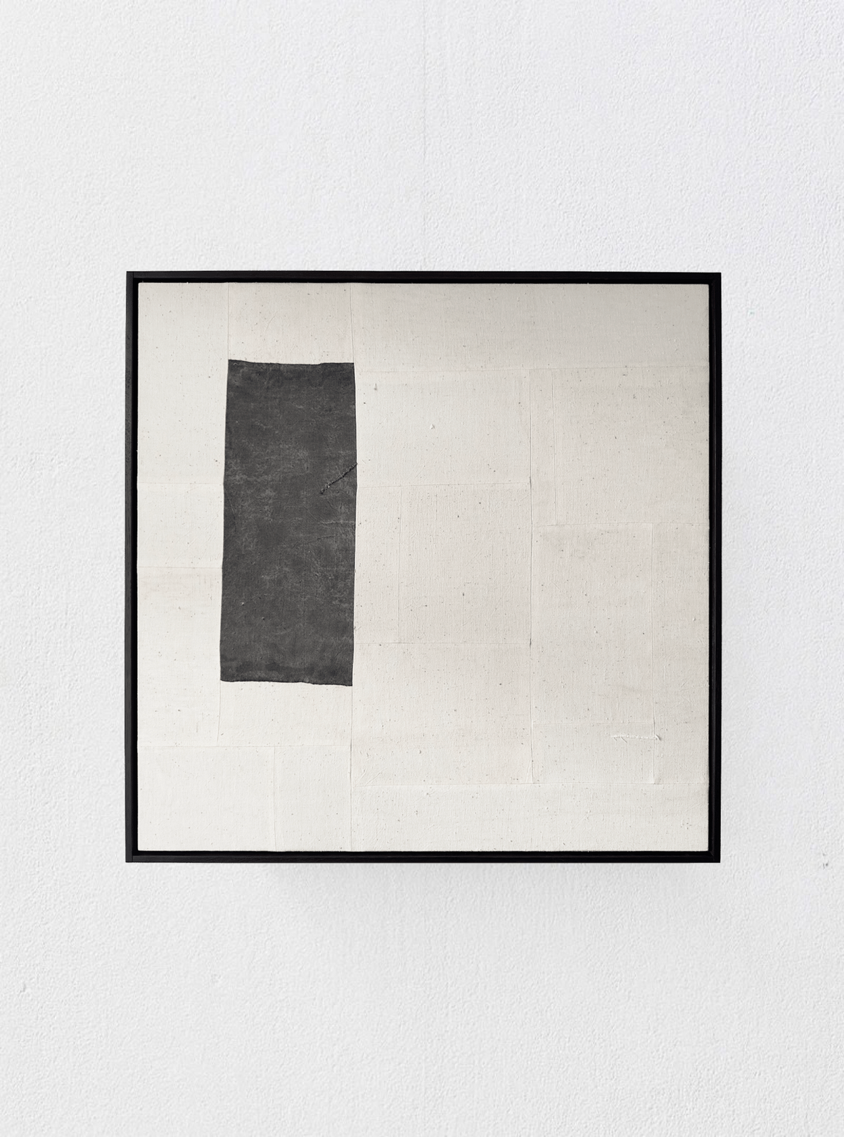A minimalist framed artwork hangs on a white wall. The piece features a large off-white canvas with a roughly rectangular-shaped, black patch in the upper left quadrant, reminiscent of traditional Chinese cotton patchwork. The art is defined by its simplicity and contrast between the neutral tones and dark element. The artwork is "(a-tent), 2024 Framed Hand Loomed Tapestry" by Bec Kirby.