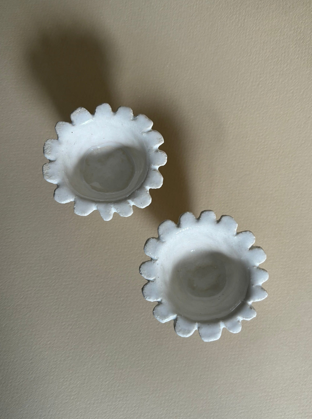 Two small, white "Rosetta" Egg Cups by Studio Beurre with scalloped edges are placed on a light beige surface. The egg cups, made from white clay, have a simple and rustic appearance, with slight variations in their shapes giving them a handmade look. Shadows fall to the left, adding depth to the image.