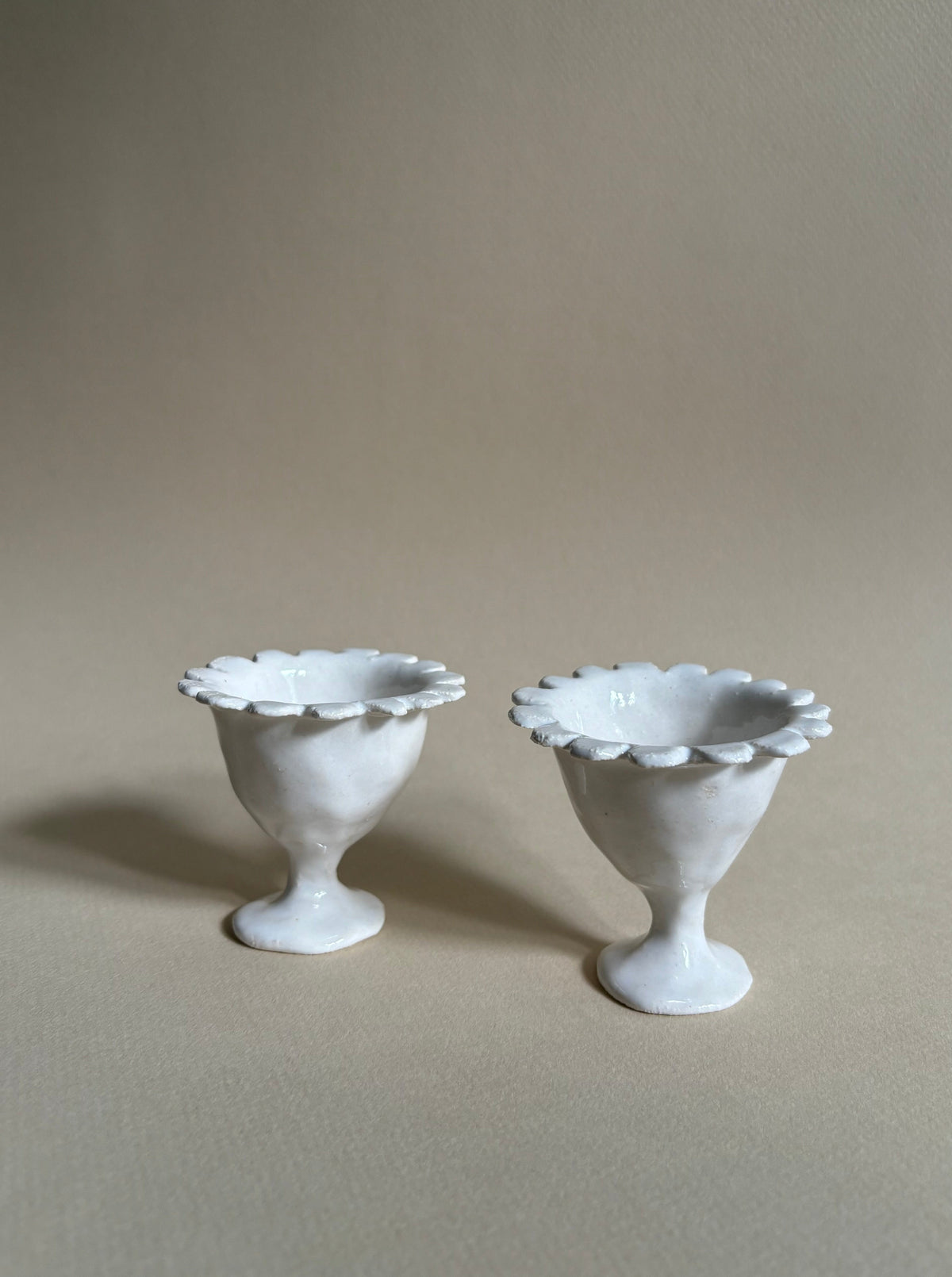A pair of the "Rosetta" Egg Cup by Studio Beurre with a fluted edge and high pedestal base, placed on a plain beige background. Made from white clay with a shiny white glaze, the egg cups boast an elegant, handcrafted appearance.