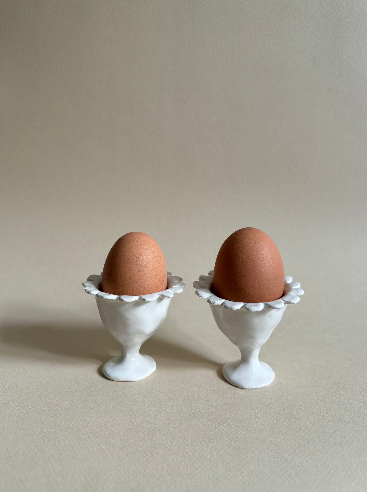 Two brown eggs are placed in white, ornate egg cups. The "Rosetta" Egg Cups, crafted by Studio Beurre from white clay with a shiny white glaze, add an artisanal touch. The background is a plain light beige, providing a simple and neutral setting that emphasizes the eggs and their holders. Soft lighting creates gentle shadows.