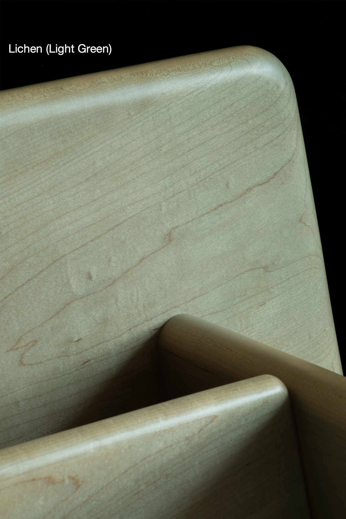 Close-up image of light green wood furniture with a smooth finish. The image highlights the corner details and the grain pattern of the wood, capturing the elegance of premium natural materials. Text in the upper left corner reads, "Lichen (Light Green) Raja Chair by Juntos Projects.