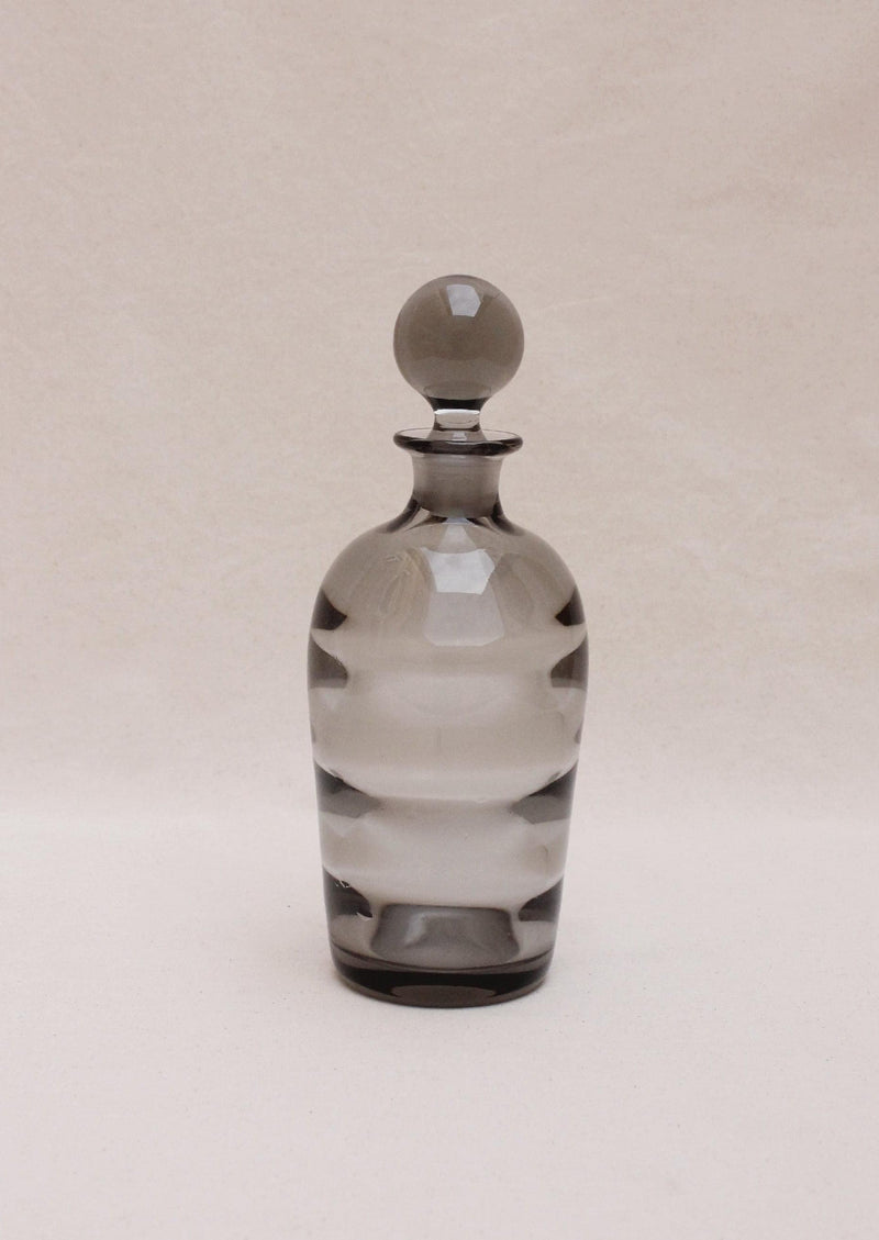 An Boga Avante Shop Art Deco liquor decanter, transparent with a rounded stopper, features alternating clear and frosted horizontal bands in an optical glass design, set against a plain, light beige background.