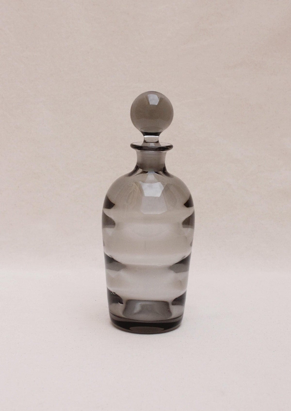 An Boga Avante Shop Art Deco liquor decanter, transparent with a rounded stopper, features alternating clear and frosted horizontal bands in an optical glass design, set against a plain, light beige background.