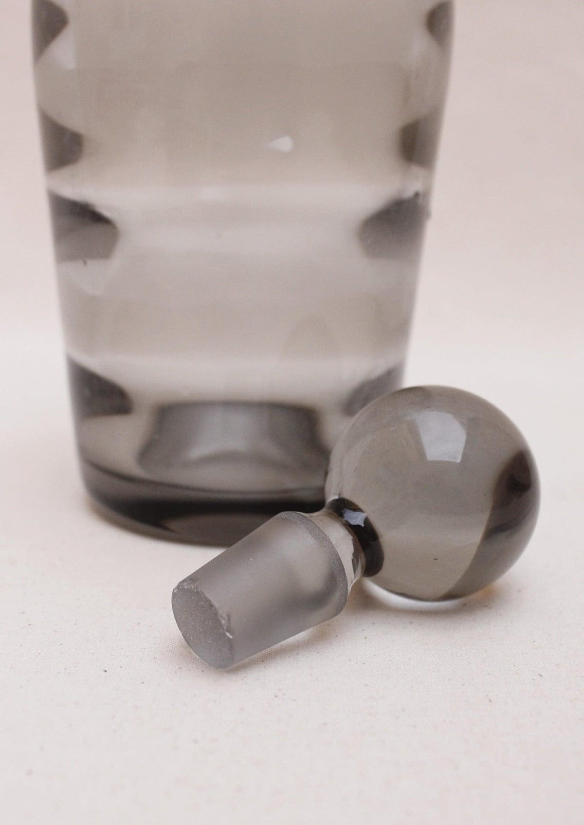 A Boga Avante Shop Art Deco Liquor Decanter with a large spherical stopper lying next to it on a textured beige surface. The glass, featuring an optical glass design, is transparent with a smoky gray tint.