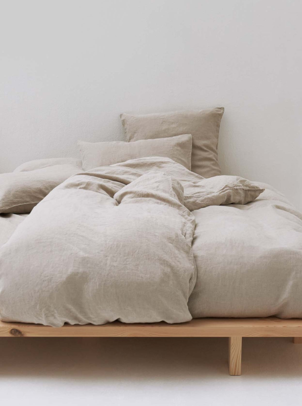 In a minimalist, well-lit room with plain white walls, the bed features the Washed Linen Bed Set in Natural by MOST and several pillows. The handcrafted wooden bed frame complements the beige duvet, enhancing the cozy and simple aesthetic with sculptural elegance.