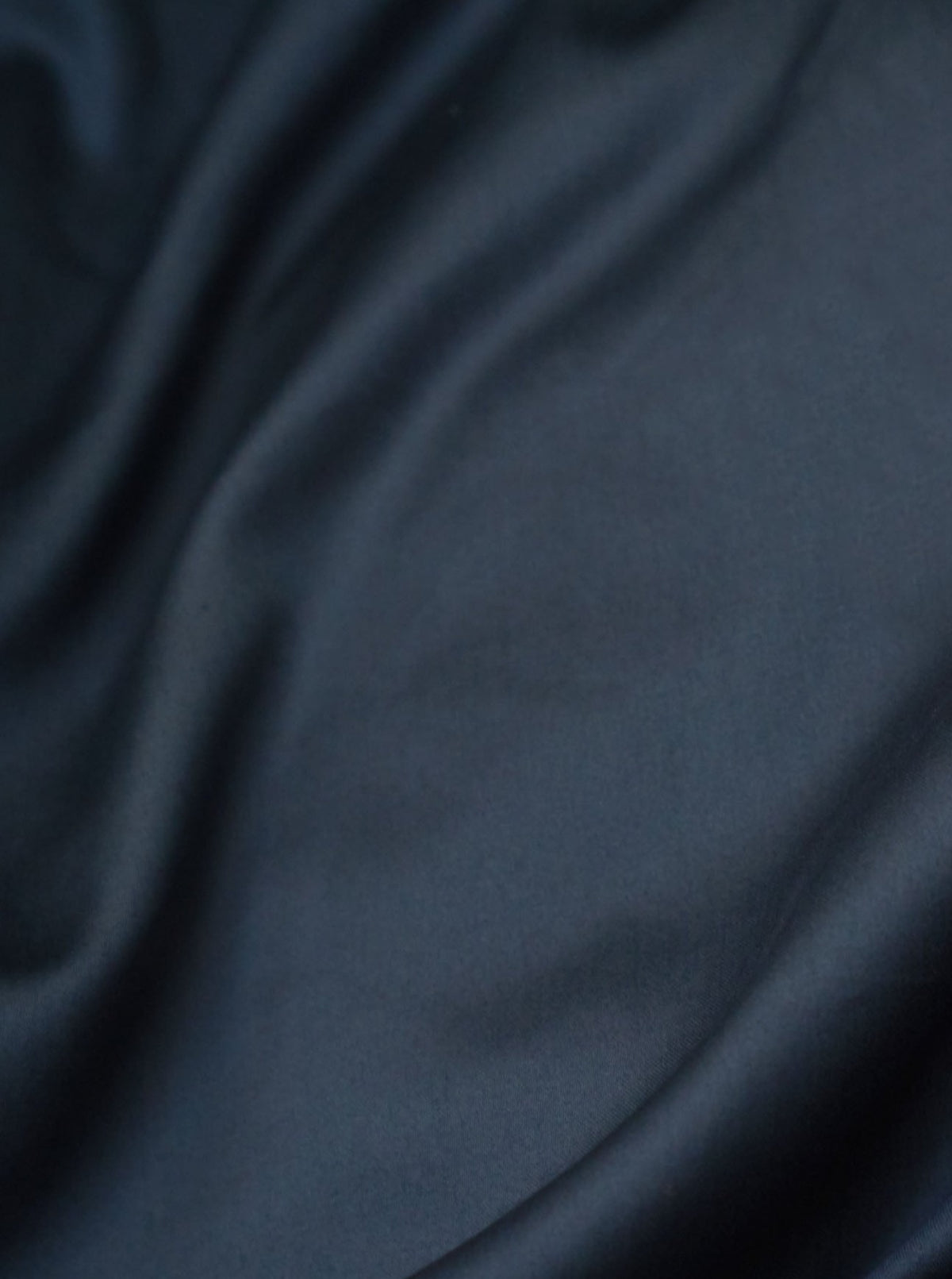 The Cotton Sateen Bed Set in Stormy from MOST features a silky dark blue hue with soft, flowing folds and a slight sheen, providing a smooth, luxurious texture perfect for high-end interior designs.