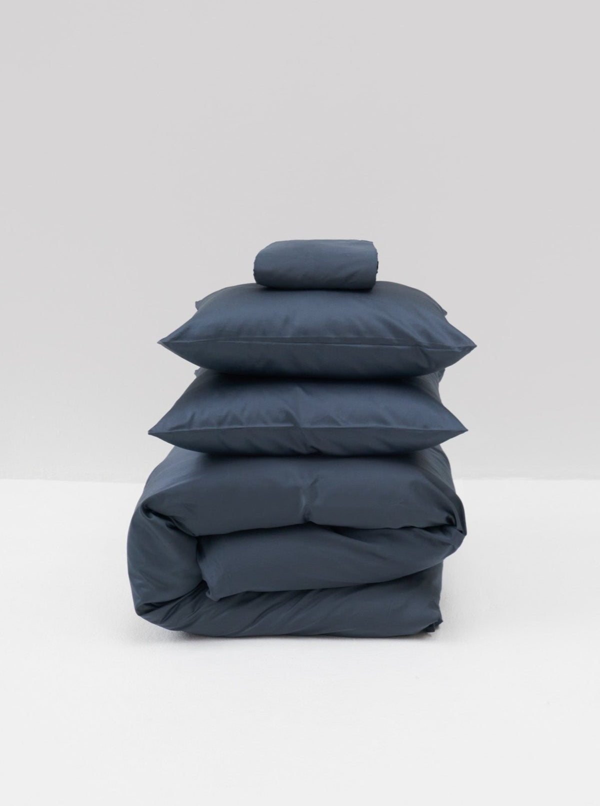 The Cotton Sateen Bed Set in Stormy by MOST, featuring a folded duvet, two pillows, and a small cushion, rests elegantly on a light gray background. This neatly arranged dark blue bedding embodies contemporary design with its luxurious touch.