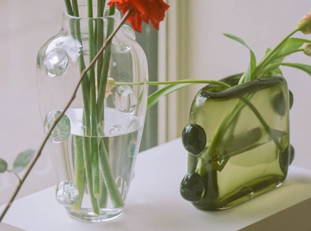 Two unique La Pierre vases on a table, one tall and clear with circular embossments, holding a flower stem from the "Asking For A Friend" series, and the other shorter, green by Sophie Lou Jacobsen.