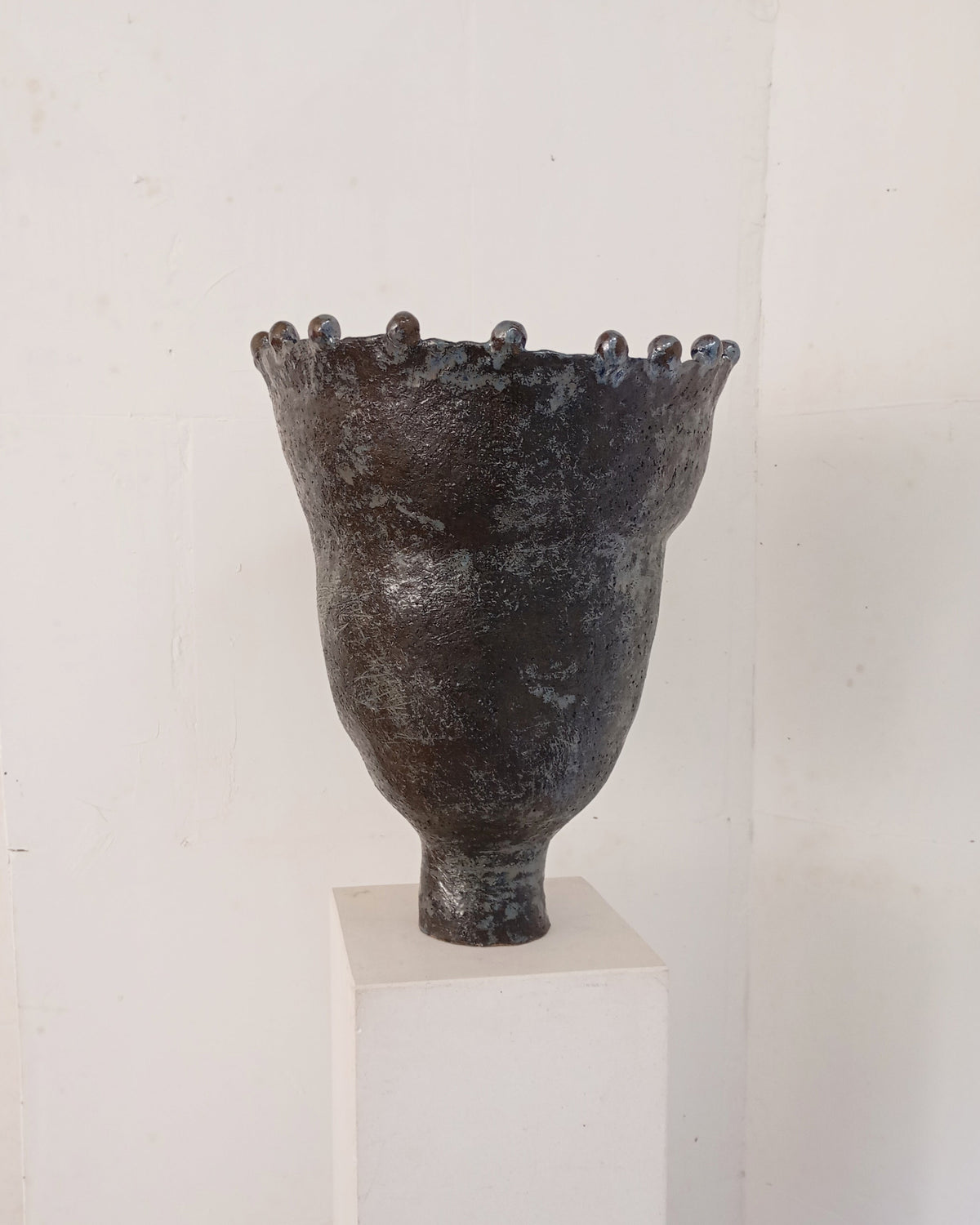 The Coupe Lilibet 7 by Claire Pain is a dark, textured ceramic vase with a narrow base, flared rim, rounded bumps along the top, and a subtle metallic glaze, shown on a white pedestal against a plain white wall.