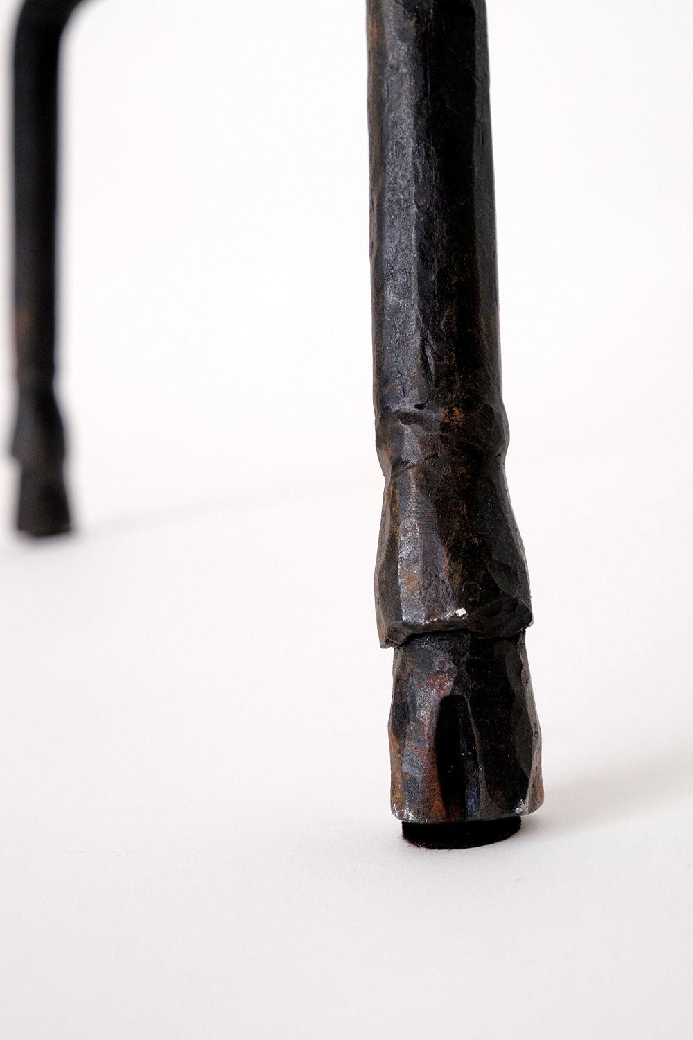 A close-up of a black, textured metal leg crafted to resemble a hoof, part of the "Sevilla" Lamp by Barracuda Interiors. Another similar leg from this zoomorphic floor lamp appears blurred in the background on a smooth white surface.