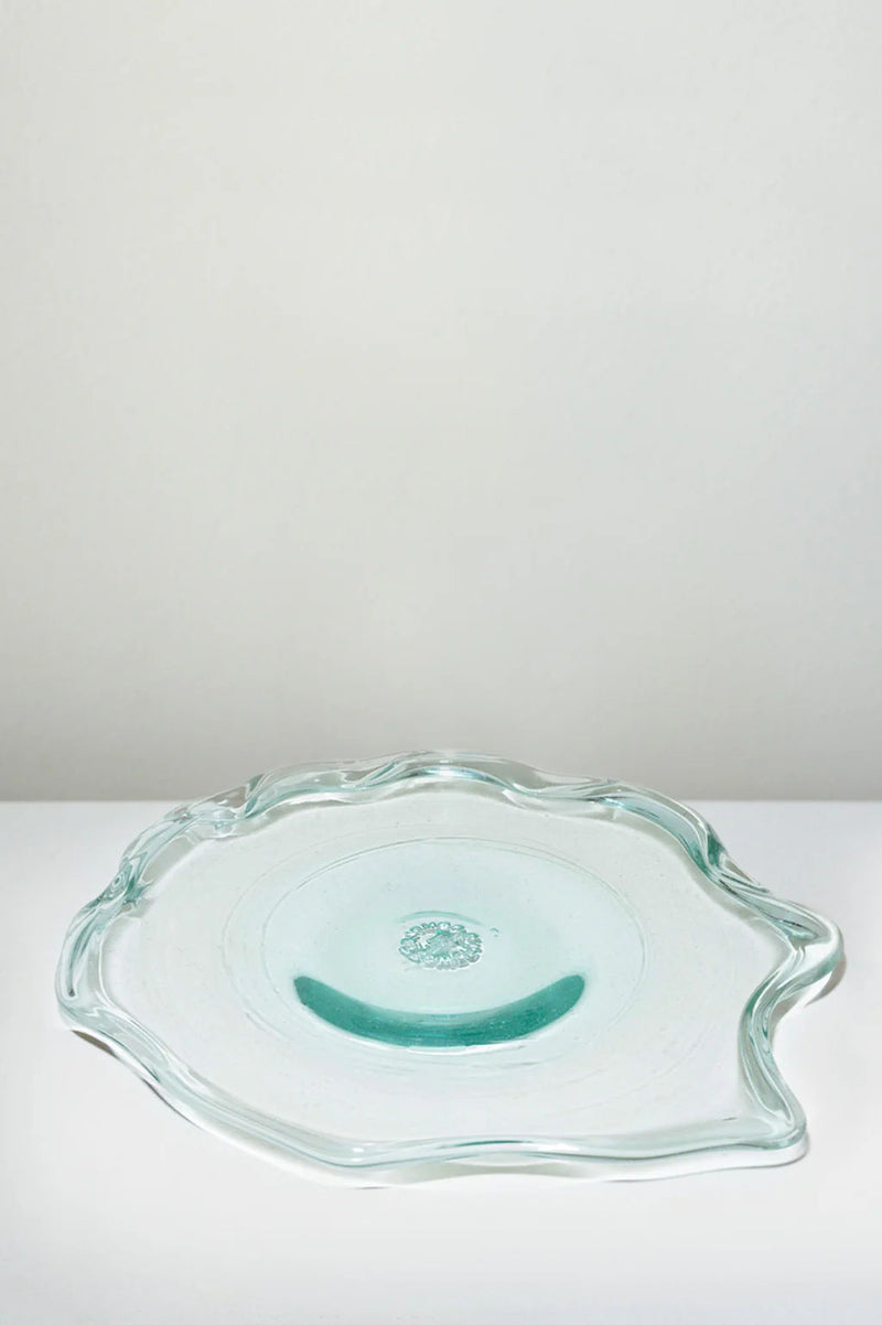 The Completedworks Pond serving plate is crafted from recycled glass with a clear, wavy-edged, irregular shape and subtle aqua tint, shown resting on a white surface against a light gray background.