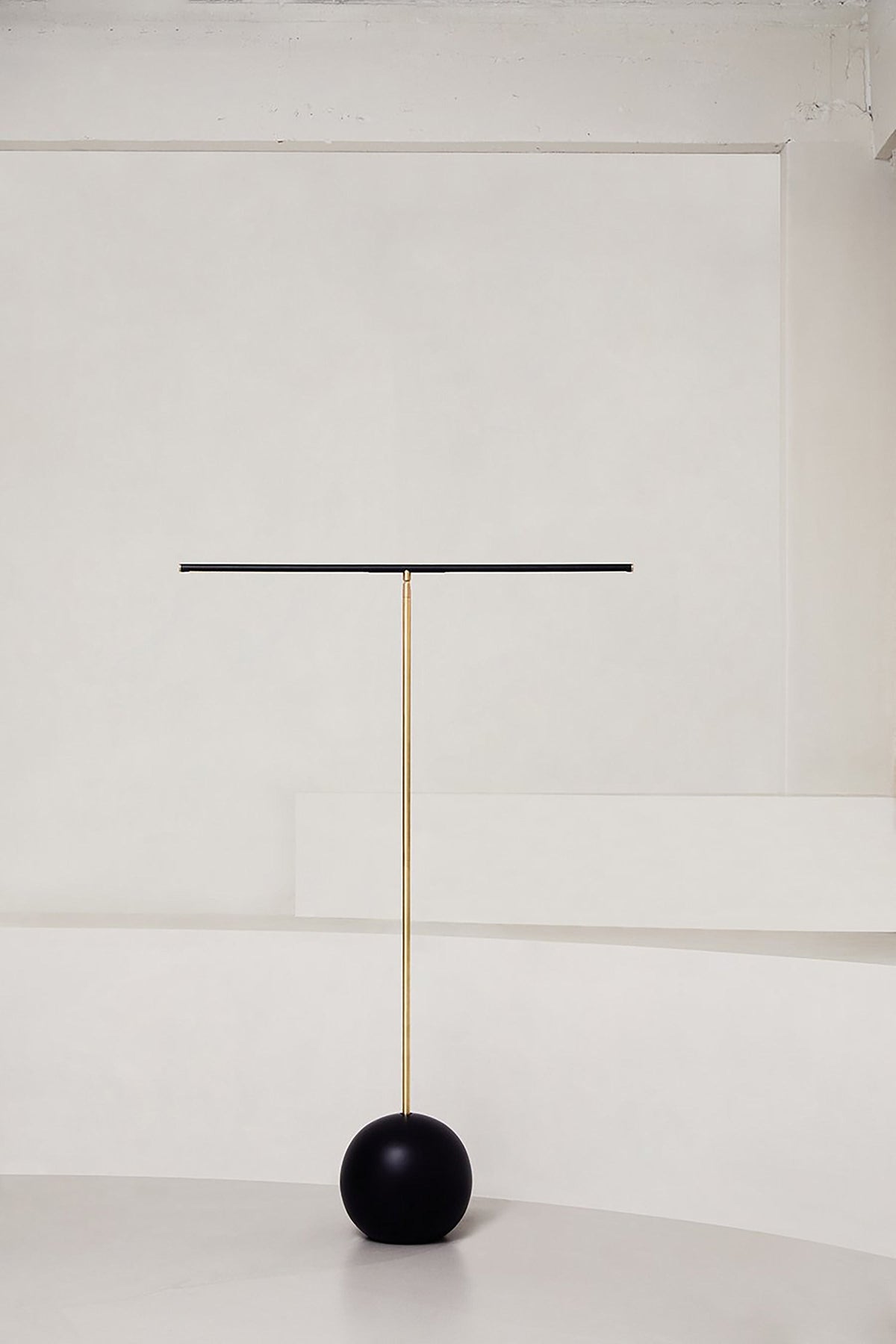 The 'T- Lamp' Floor Lamp by Heako Studio features a thin horizontal black bar on a slender brass pole with a round black base, offering minimalist, sculptural lighting against a plain white background.