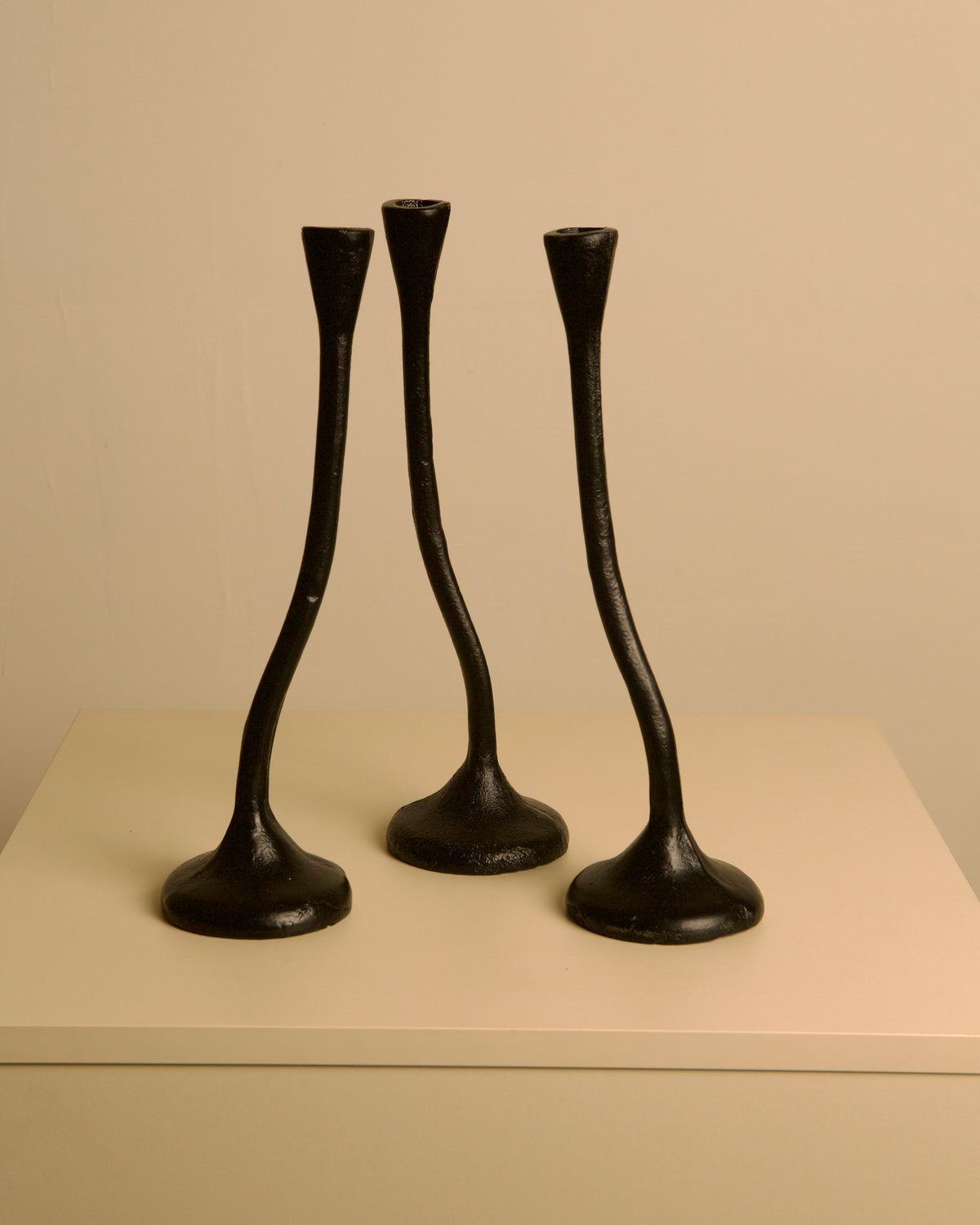 The Set of 5 "Twisted" Candleholders by Treaptyque features black cast aluminum stems with curving, sculptural lines inspired by 80s design, displayed on a light minimalist surface against a plain beige background.