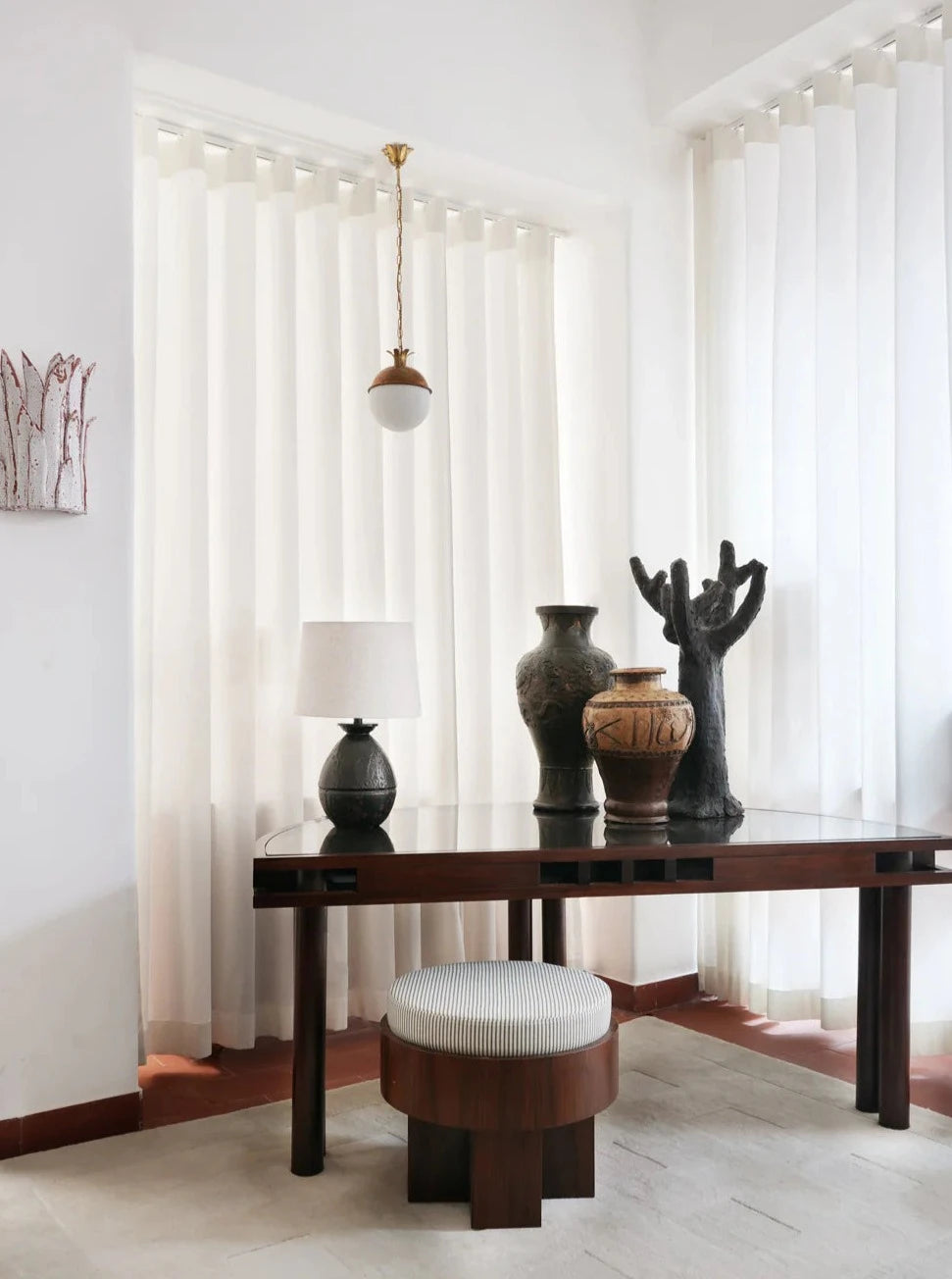 A modern interior includes a wooden table with three sculptural vases, a black lamp, and a round upholstered stool. Sheer white curtains and the Barracuda Interiors Granada Ceiling Pendant Lamp finish the minimalist, elegant space.