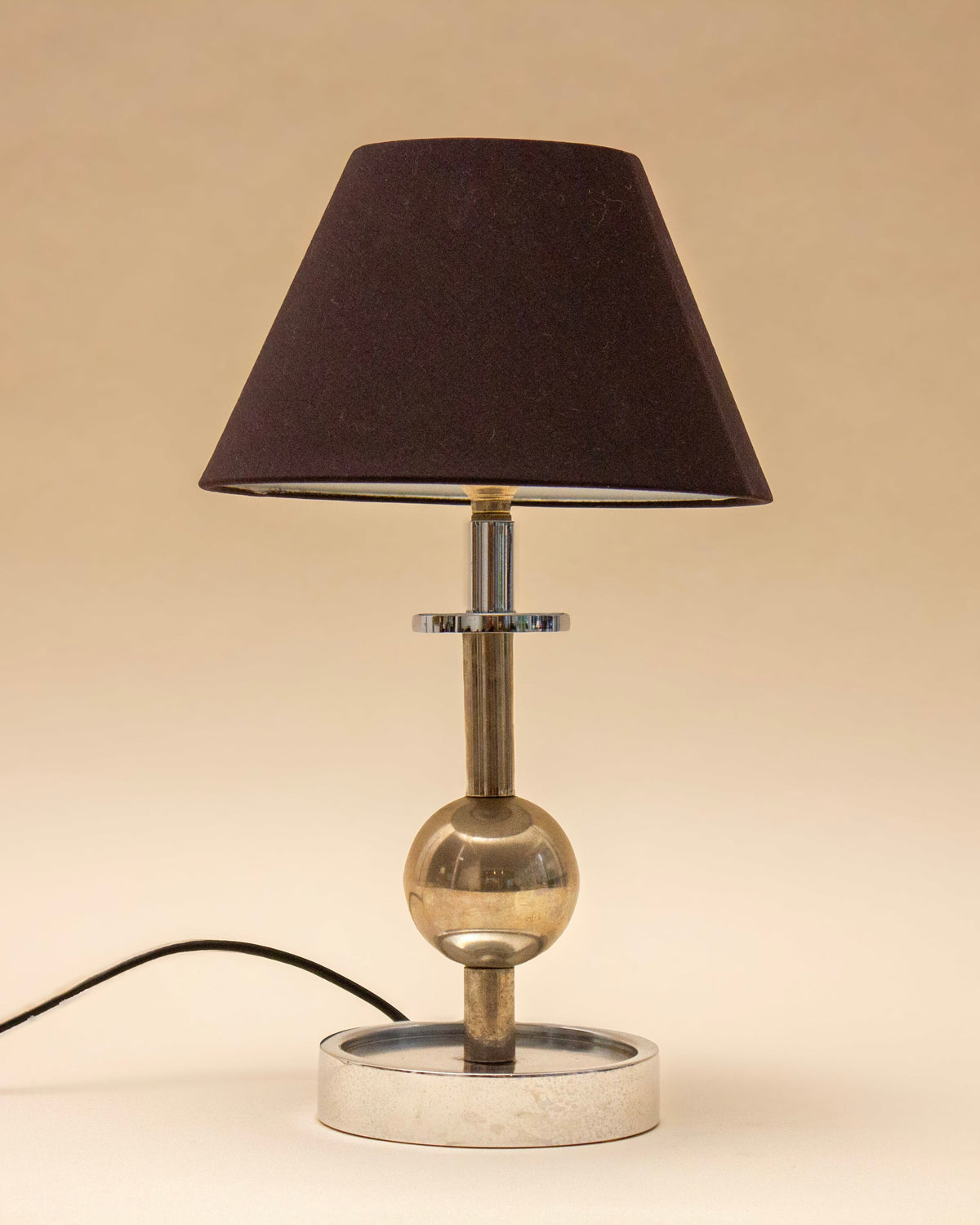 The SÒLER ALTEA TABLE LAMP features a black lampshade, metallic base, and round central accent. It stands on a light surface with its power cord visible, set against a beige background.