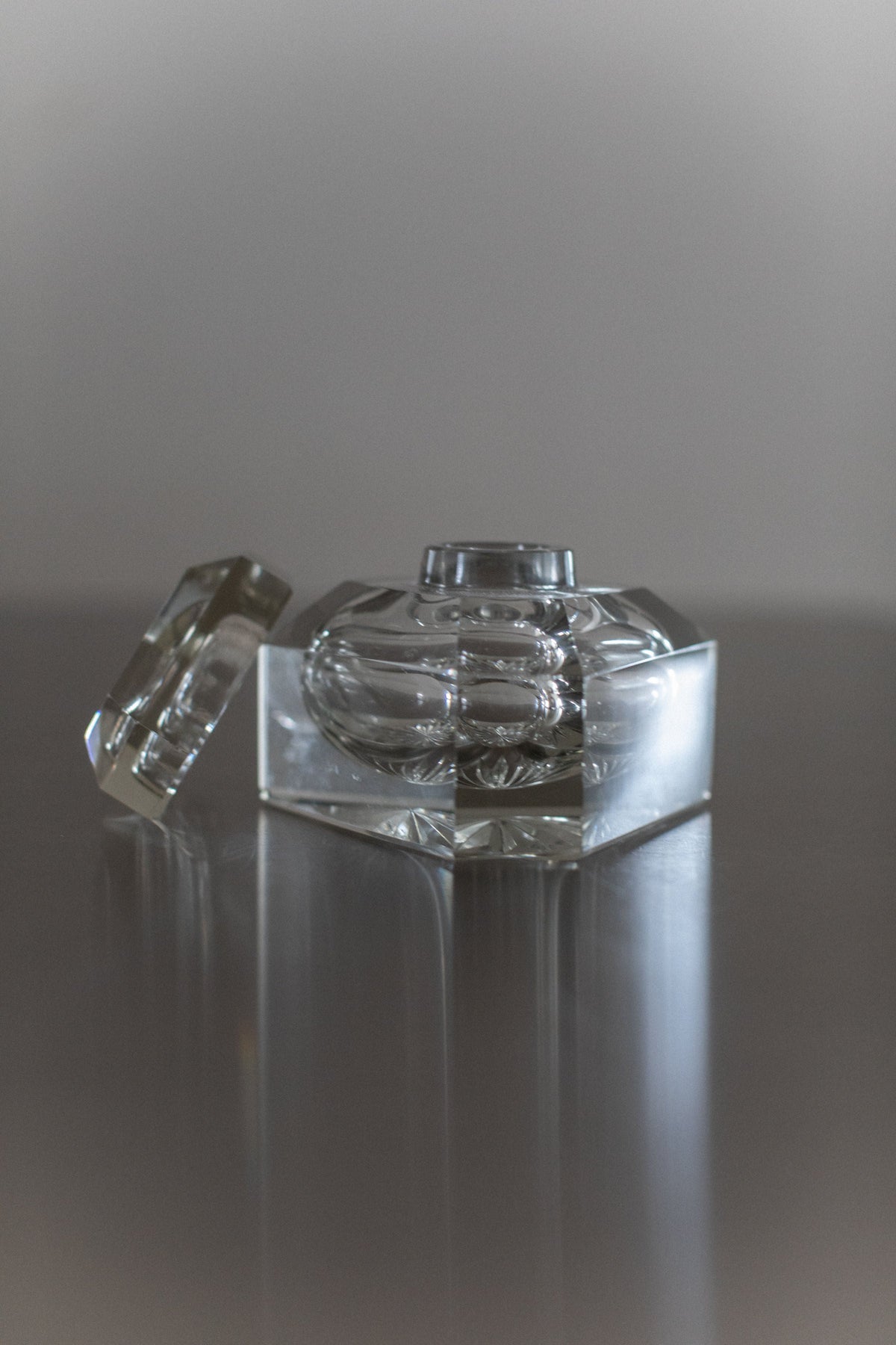 A hexagonal Glass Perfume Bottle by Out For Lunch, featuring hand-painted details and a matching lid set askew beside it, both displayed on a reflective surface with a softly blurred neutral background.
