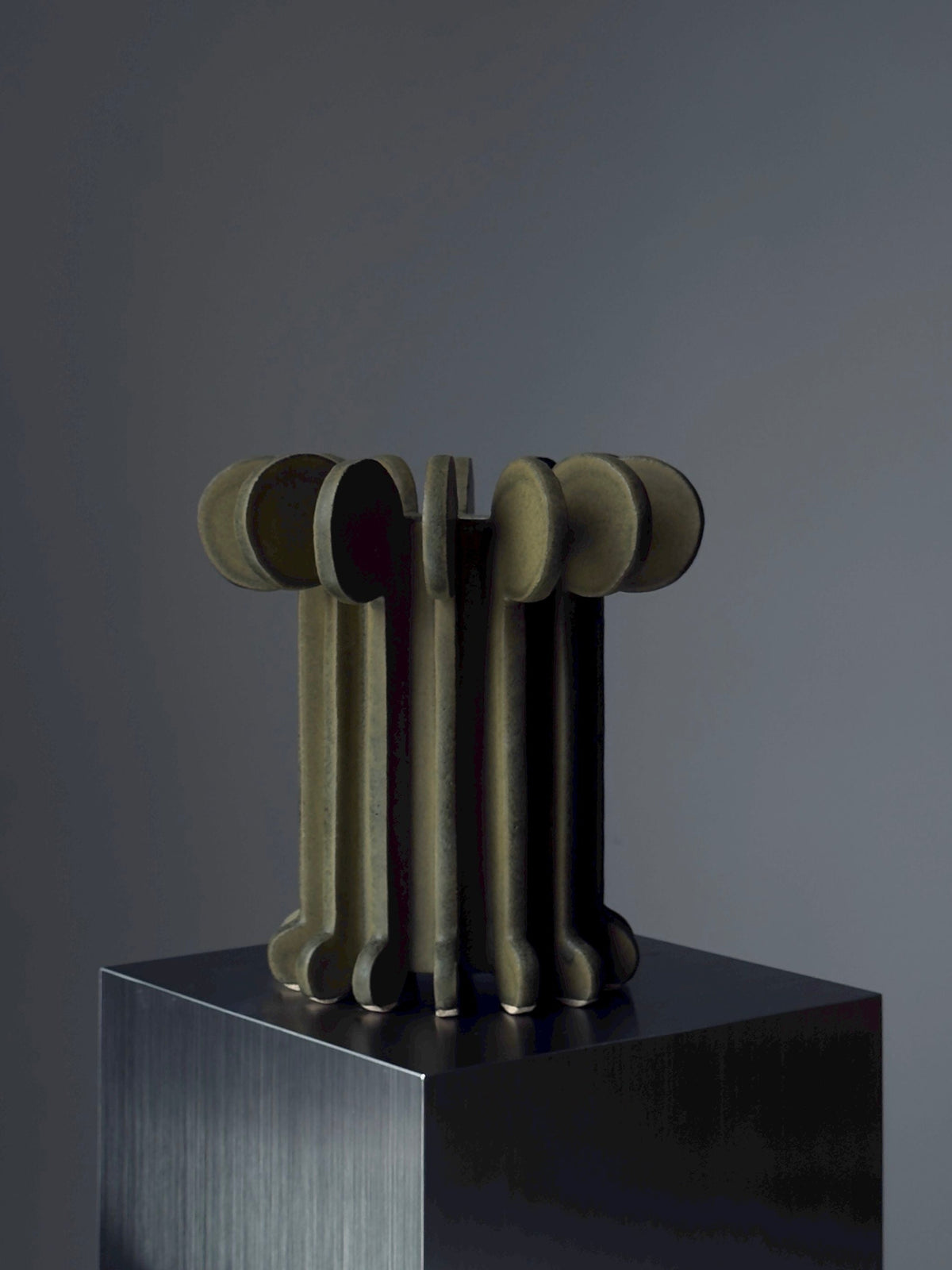 Vessel No. IV by Devin Wilde is a sculptural artwork featuring vertical, cylindrical forms with rounded ends, clustered on a metallic pedestal against a dark, neutral background.