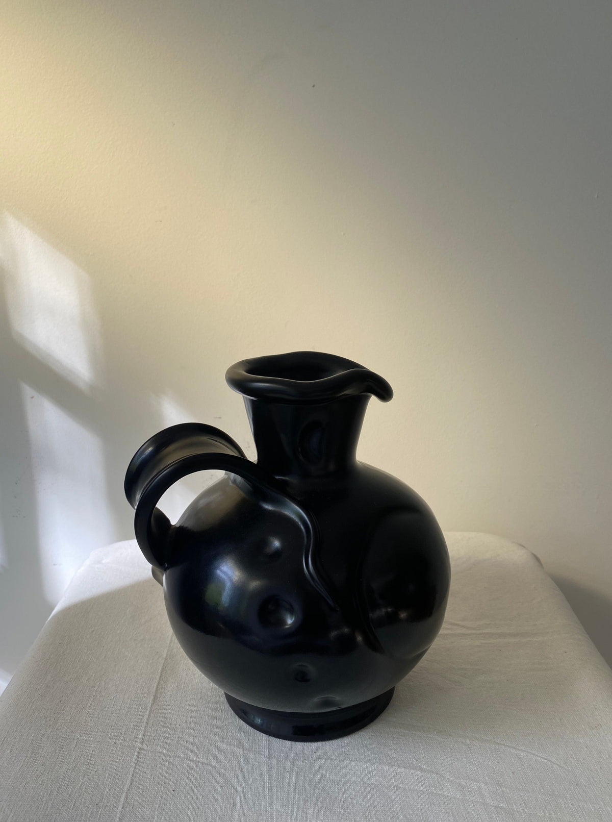 The Art Deco Vase by Les Objets Amsterdam features a glossy black ceramic finish, round body, curved handle, and sculpted bumps, exuding elegance as it rests on a white cloth against a softly lit neutral wall.