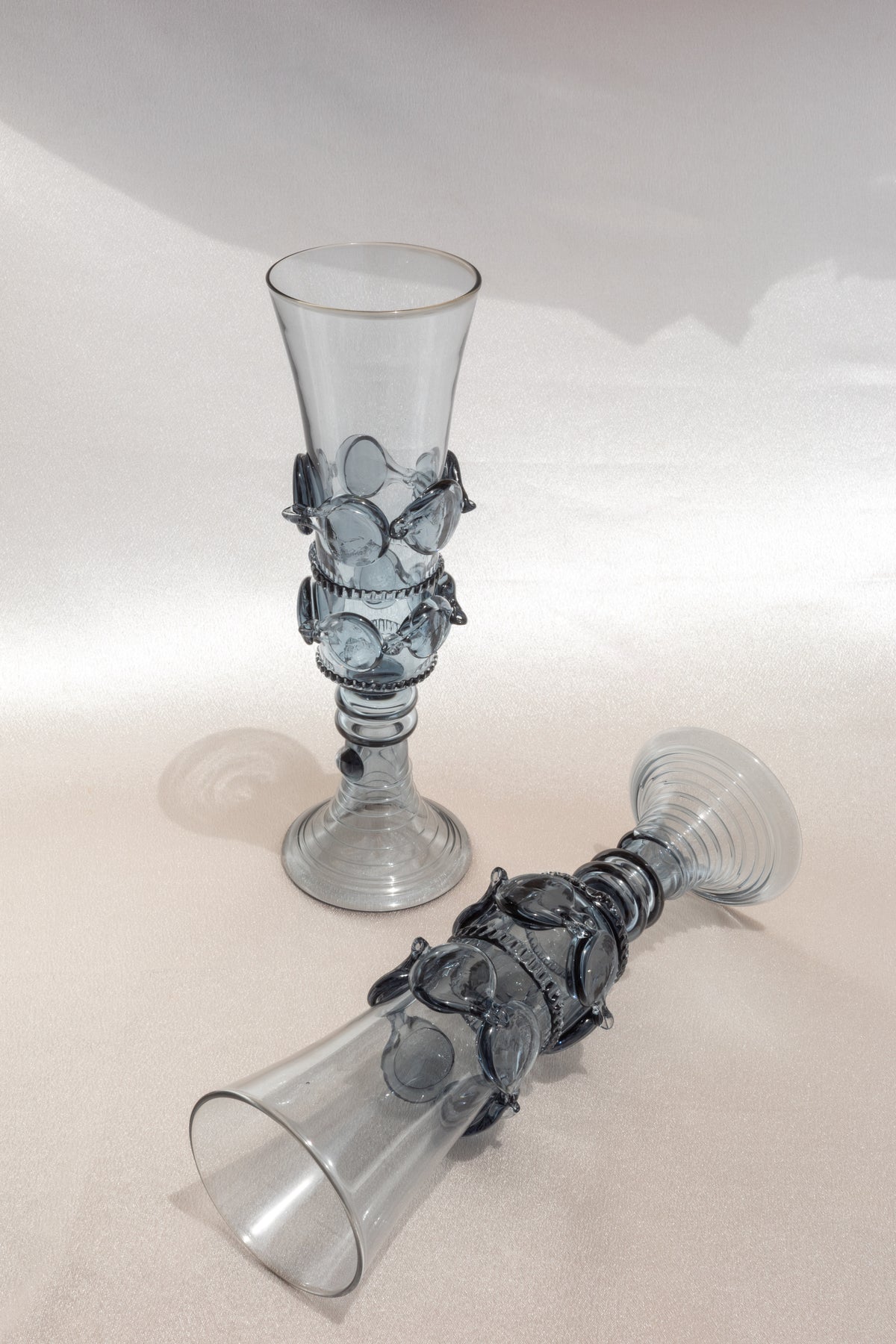 A pair of elegant, translucent Lucie Claudia Podrabska mouthblown champagne flutes from the Dots Spirals collection—one upright, one on its side—rest on a light textured surface.