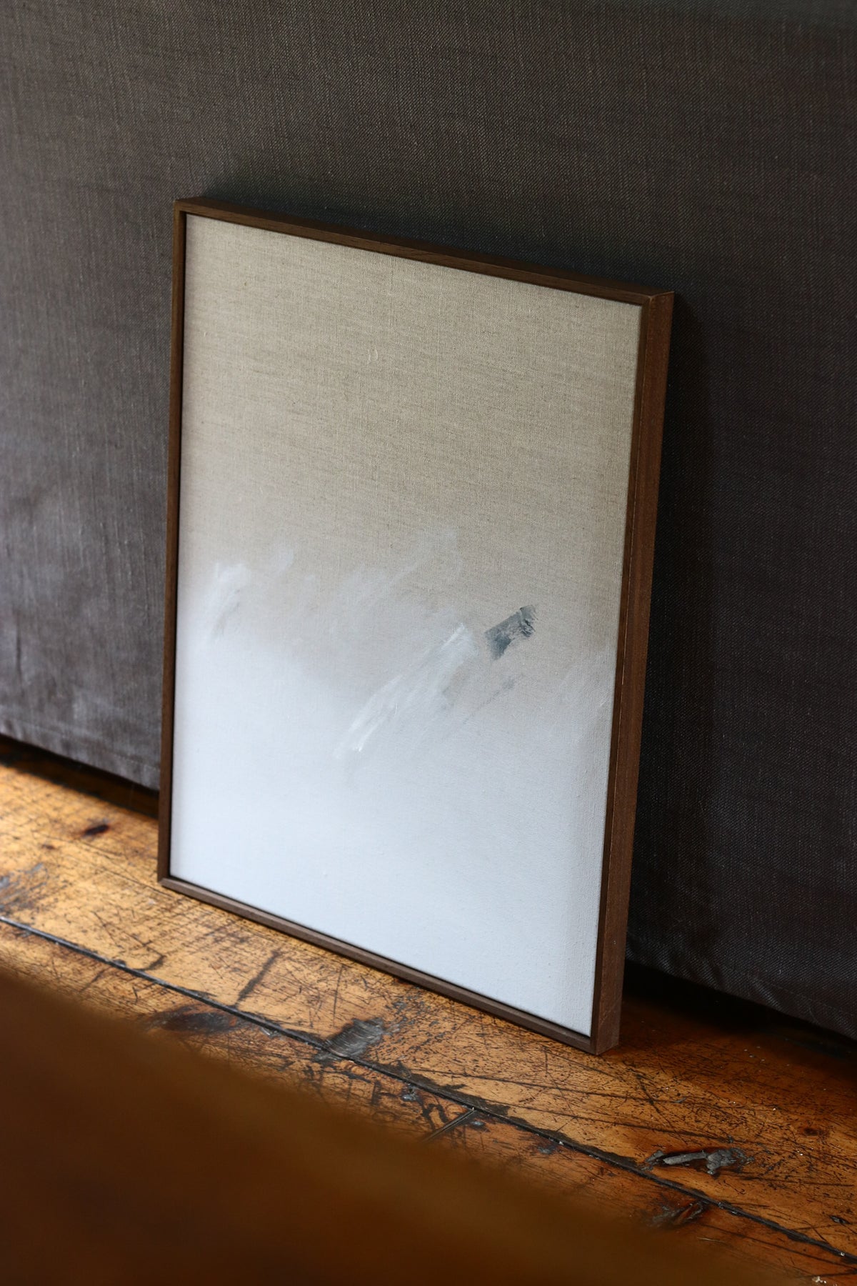 Holding — Acrylic on Linen by David Hardy, a minimalist modern painting with soft white and gray brushstrokes, leans against a dark fabric wall on a wooden floor.