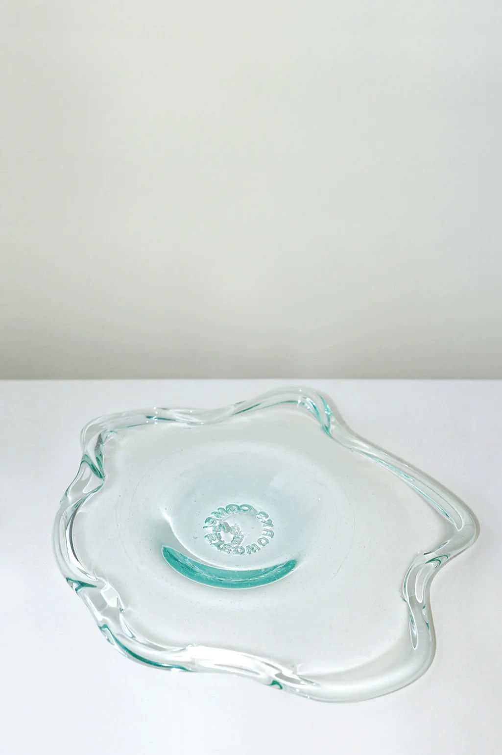 The Pond - Recycled Glass Serving Plate in Clear by Completedworks is an irregularly shaped dish with wavy edges, crafted from recycled glass and displayed on a white surface against an off-white background.