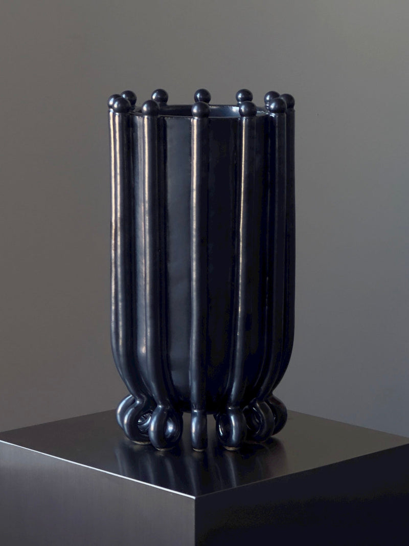 Vessel No. III by Devin Wilde is a tall, cylindrical black vase featuring vertical ridges and rounded bumps at the top and bottom, shown on a dark pedestal against a neutral background.