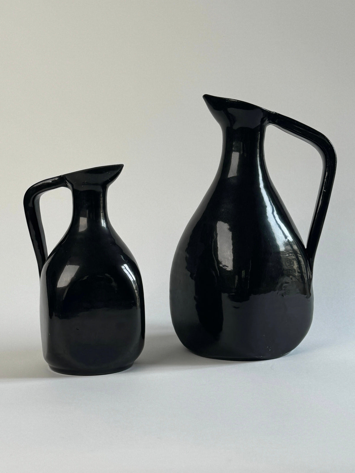 A Pair of Accolay Ceramic Carafes by Figuresdesign, featuring glossy black curved bodies and handles—one large, one small—are displayed side by side against a neutral light gray background.