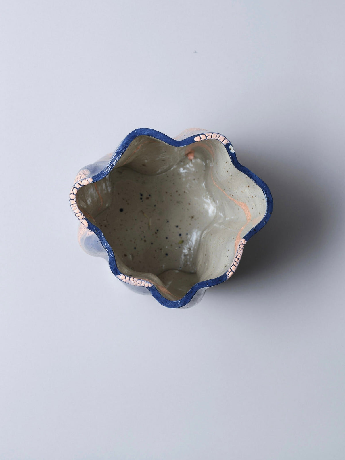 Top view of Astrid Öhman’s Swirly Vase Blue and Peach: a handmade stoneware vase with a wavy, dark blue rim accented by pink cracks and a light speckled glaze lining the interior, set against a plain background.