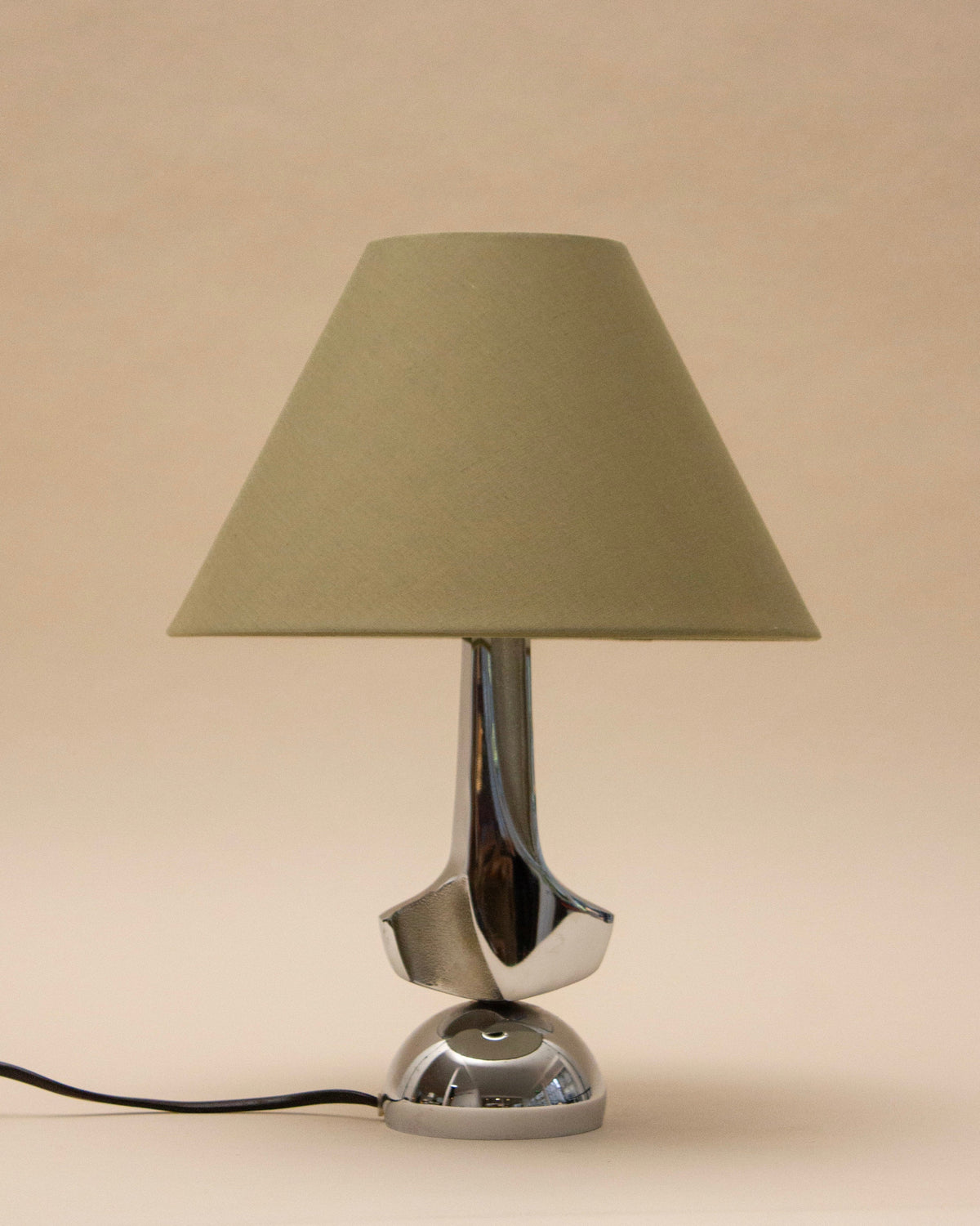 The SÒLER MELLILA TABLE LAMP features a modern chrome design with a beige conical shade and shiny silver base, set against a plain beige background. A black power cord extends from the base.
