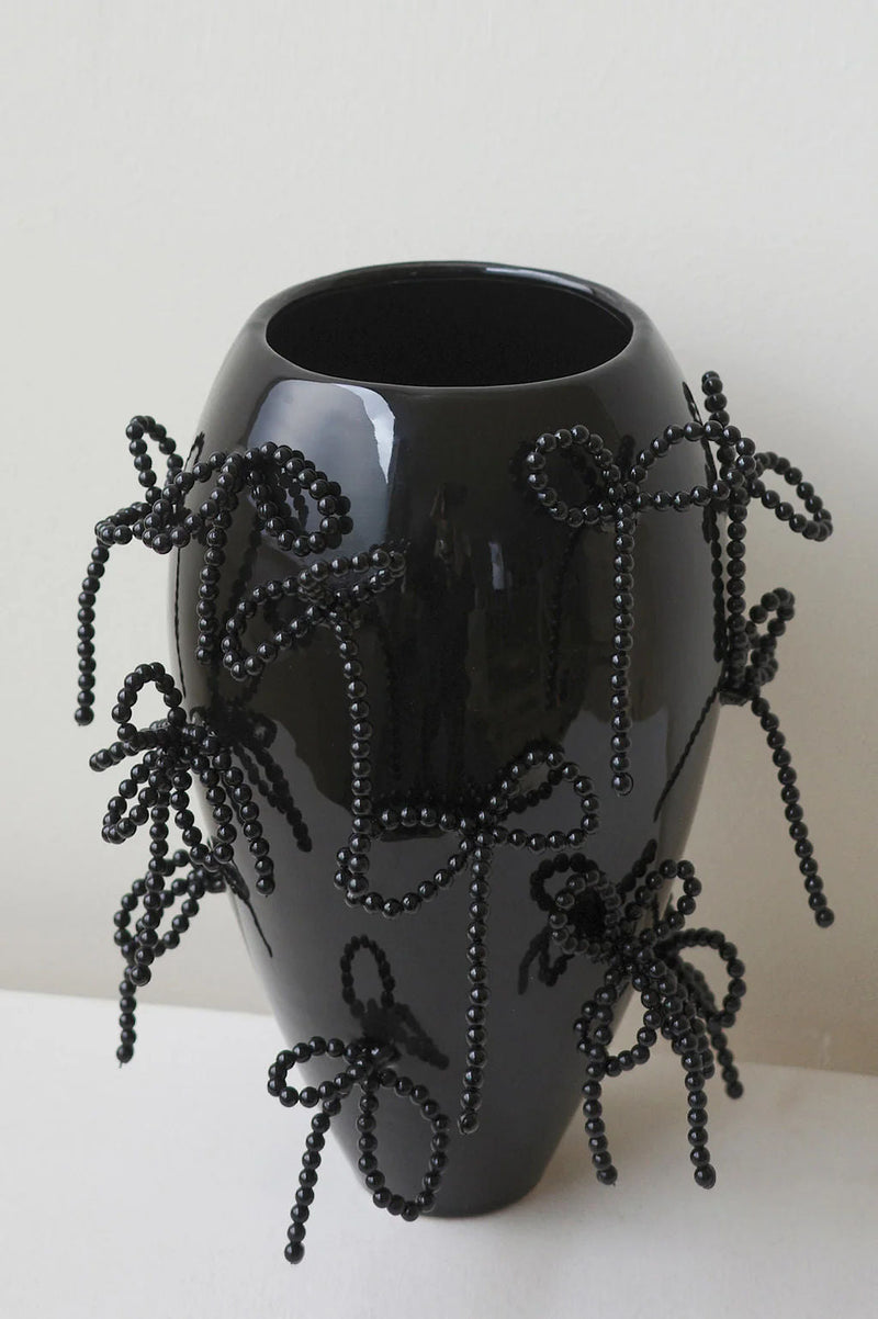 The Pearly Pearl - Large Vase In Gloss Black by Completedworks features glossy black ceramic adorned with black beaded bows, embodying conceptual ceramics. It stands on a light surface against a plain background.