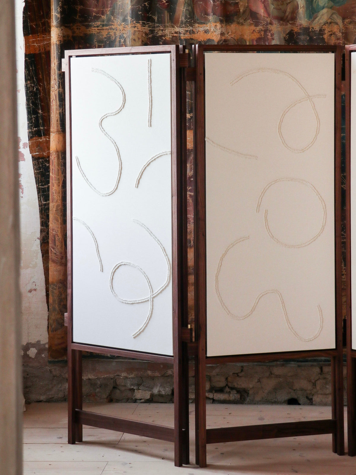 The Cavalletto Room Divider by Filippo Andrighetto, with solid walnut and white panels featuring abstract curved lines, stands on a light wooden floor before a textured wall with peeling paint and a decorative mural.
