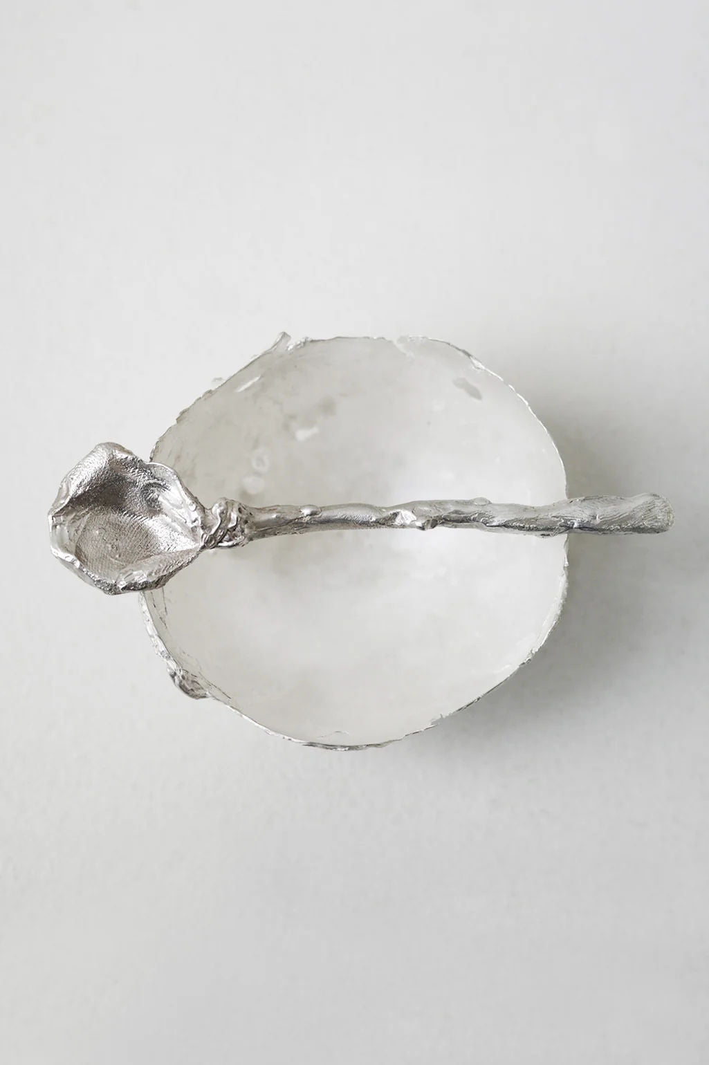 The L32 – Spoon in Silver by Completedworks rests across a handcrafted silver bowl, both showcasing a rough, organic texture and sculptural forms, set against a light gray background.
