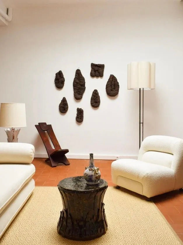 A modern living room with Barracuda Interiors' West African Wood Chair (1960s), white armchairs, two lamps, a textured black coffee table, dark organic wall art, terracotta tiles, and a beige rug for warmth.