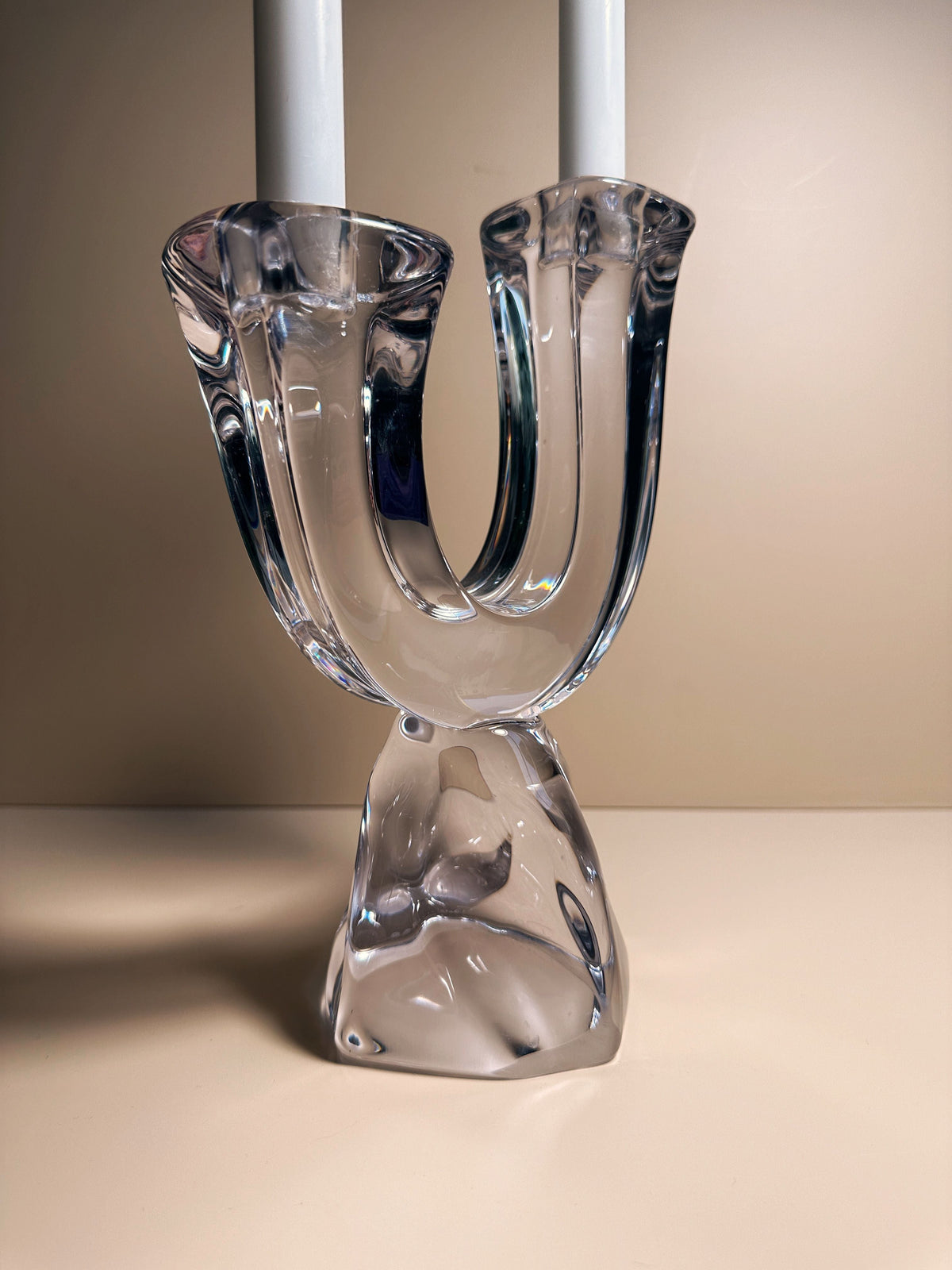 The Solid Crystal Glass Brutalist Candelabra by Daum (1950s) from Dodo Vintage features two upward-curving arms with white candles and a bold, artistic base, shown on a beige surface against a matching background.
