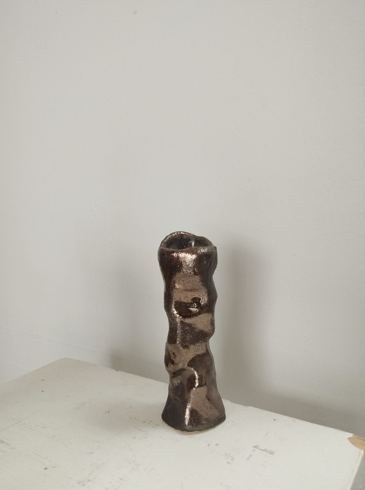 A small, metallic ceramic vase with a bronze enamel finish sits on a white surface beside the Claire Pain Candleholder in sandstone and bronze enamel, creating an eye-catching display with organic shapes and shimmering accents.