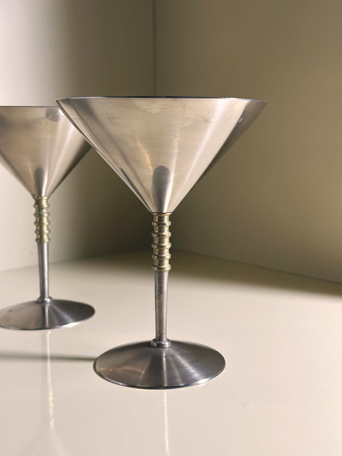 Two solid metal coupés with brass details from Dodo Vintage are placed on a white surface. One glass (12.5cm high) stands in the foreground, while the other is partially visible behind it against a light background.