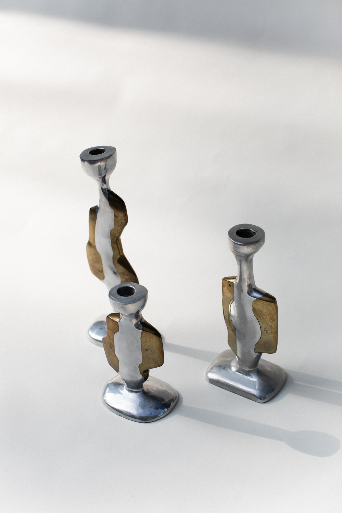 Three Brutalist Candleholders by Out For Lunch—modern metallic designs with abstract silver and brass elements—are arranged on a smooth, light surface with soft natural light and shadows.