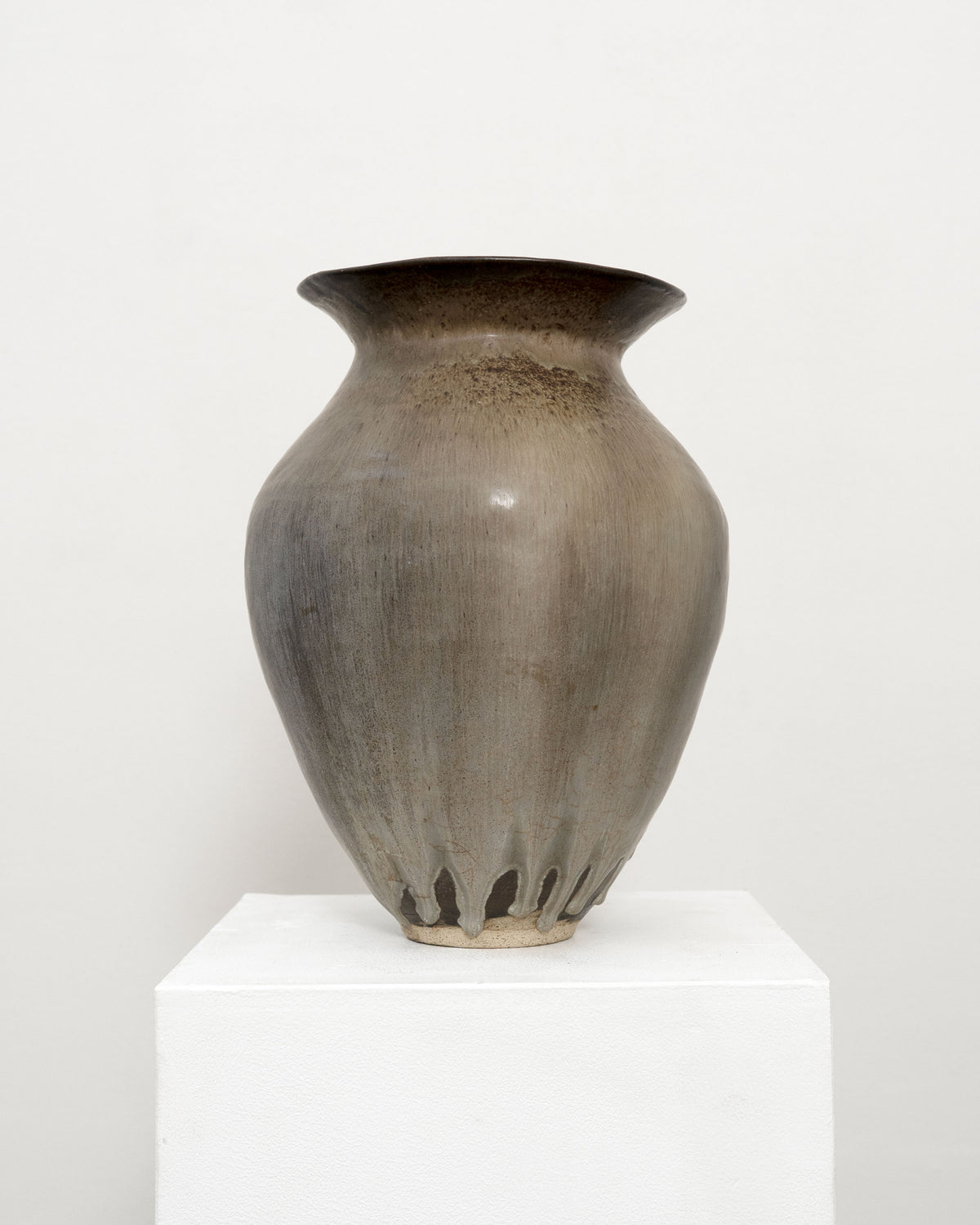 The Hand Built Studio Pottery Vase by Tom Bogle is a large, smooth brown stoneware piece with a flared rim, subtle vertical grooves, and unique irregular cut-outs at the base. It sits on a white pedestal against a plain background.