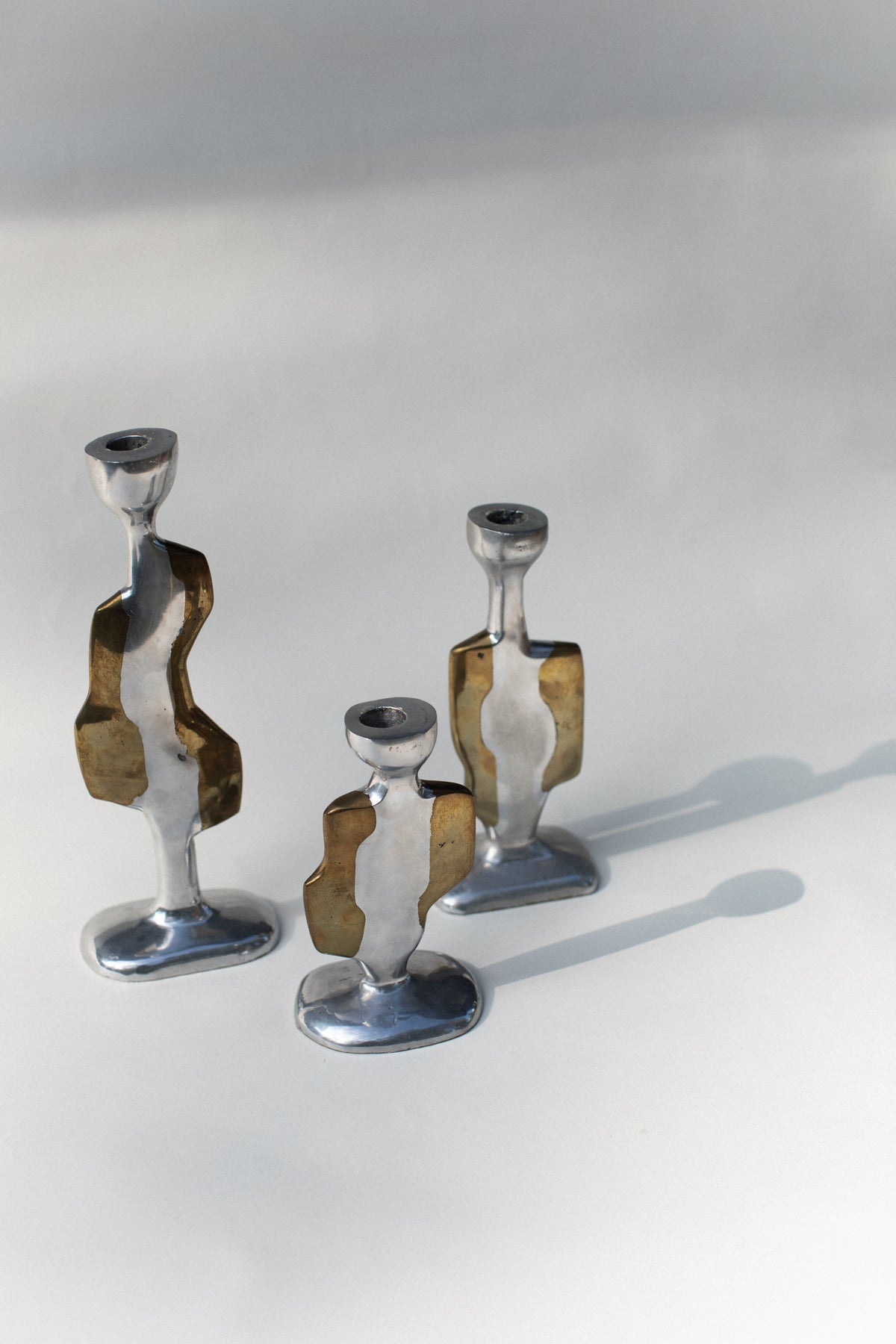 Out For Lunch’s Three Brutalist Candleholders, in varying heights with abstract curves and mixed silver and brass tones, stand on a light surface casting soft shadows. Inspired by David Marshall Disenos, the background is minimal and softly lit.