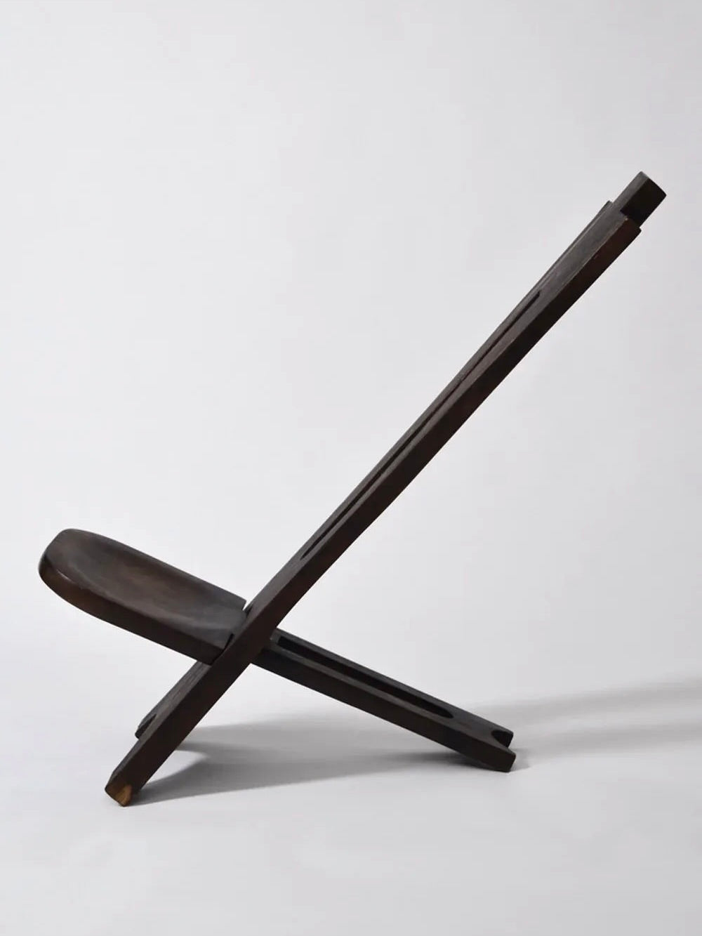 Side view of the Barracuda Interiors West African Wood Chair, 1960s—a minimalist dark wooden folding chair with a low seat and angled backrest—stands out against a plain white background.